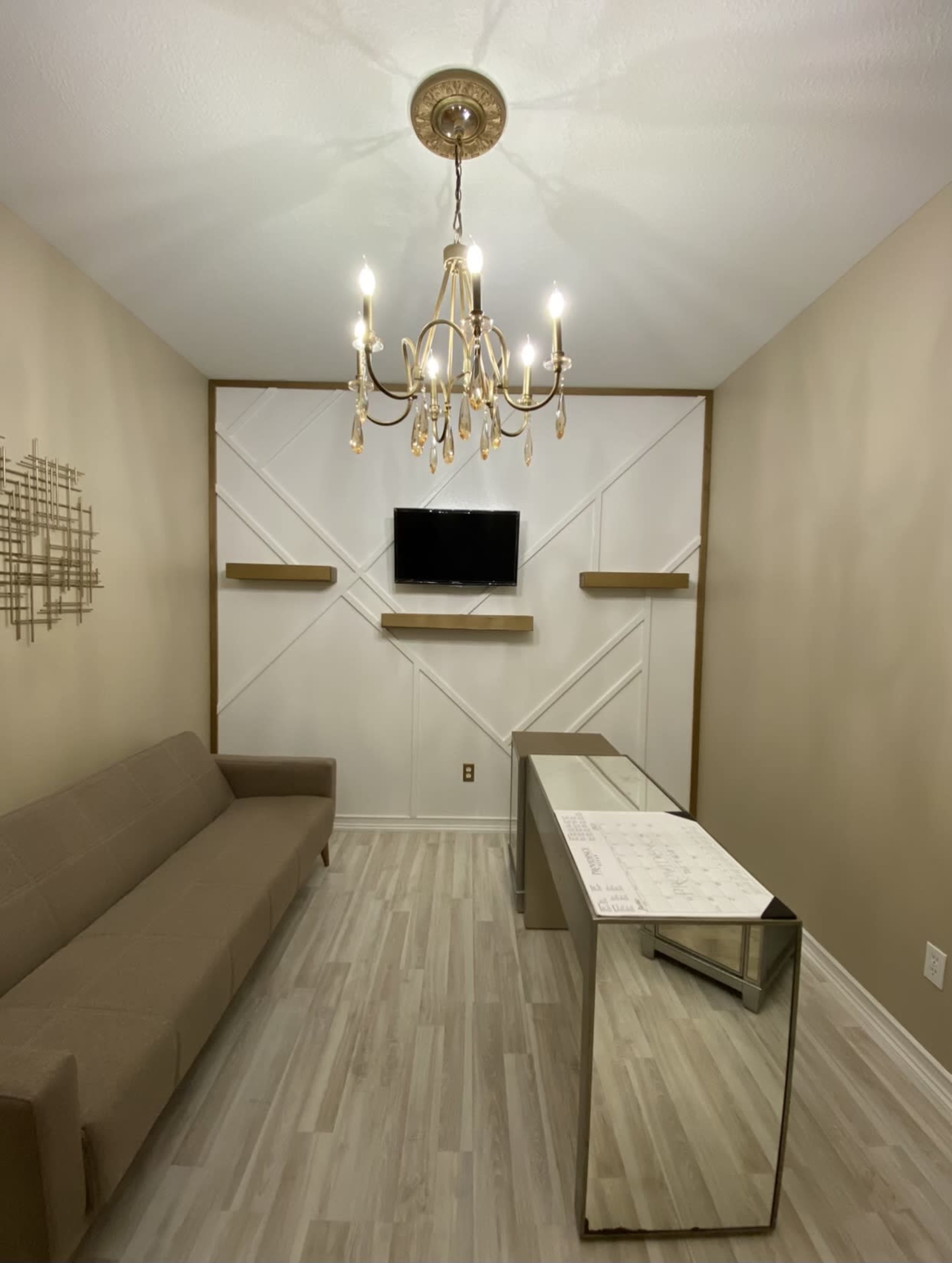 A small, elegantly designed room featuring a beige sofa, a glass desk, a chandelier, and a television mounted on the wall.