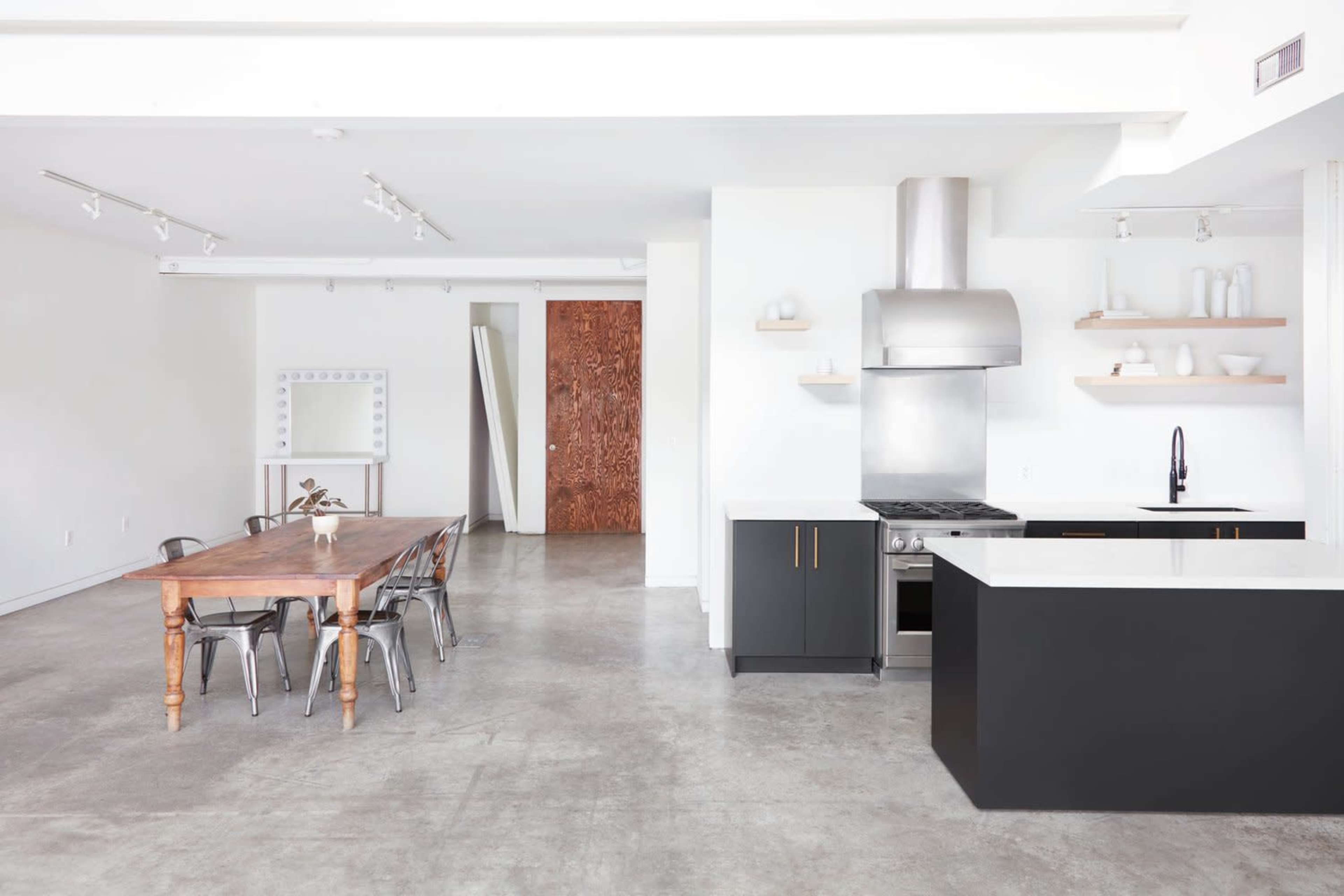 A spacious kitchen and dining area with a long wooden table, black cabinetry, a stainless steel stove, and concrete flooring.