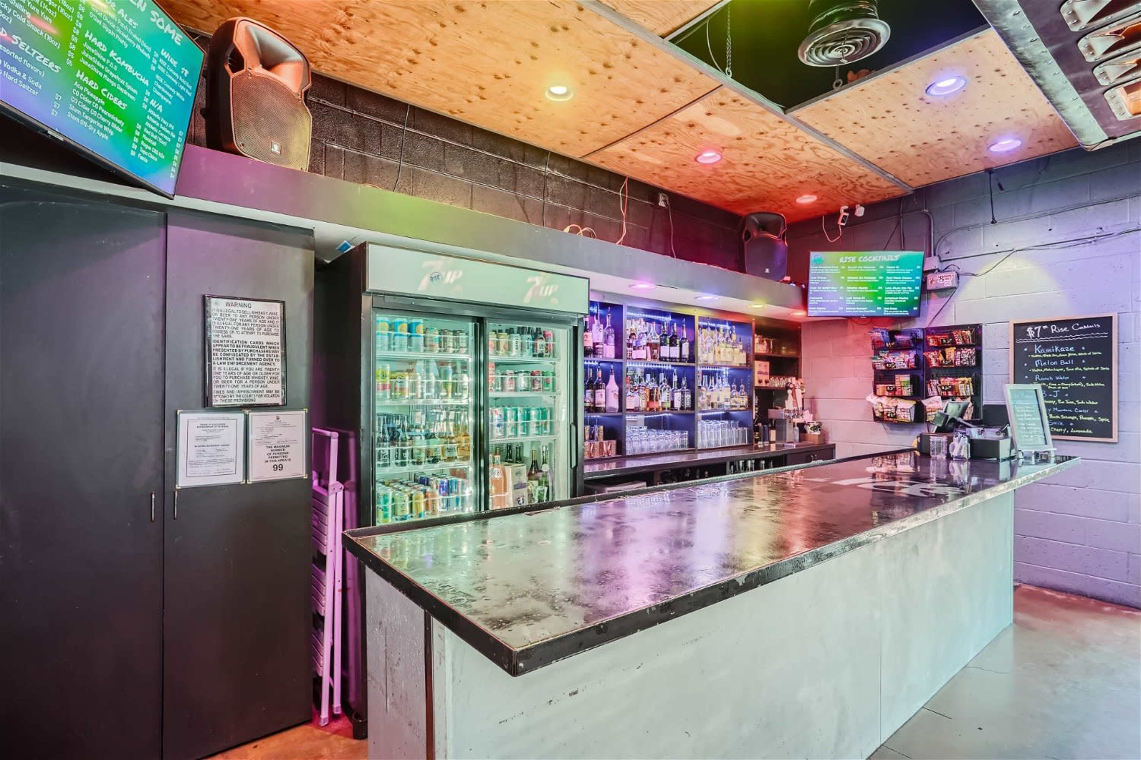 A modern bar with a concrete countertop, colorful lighting, and shelves displaying various bottles of alcoholic beverages.