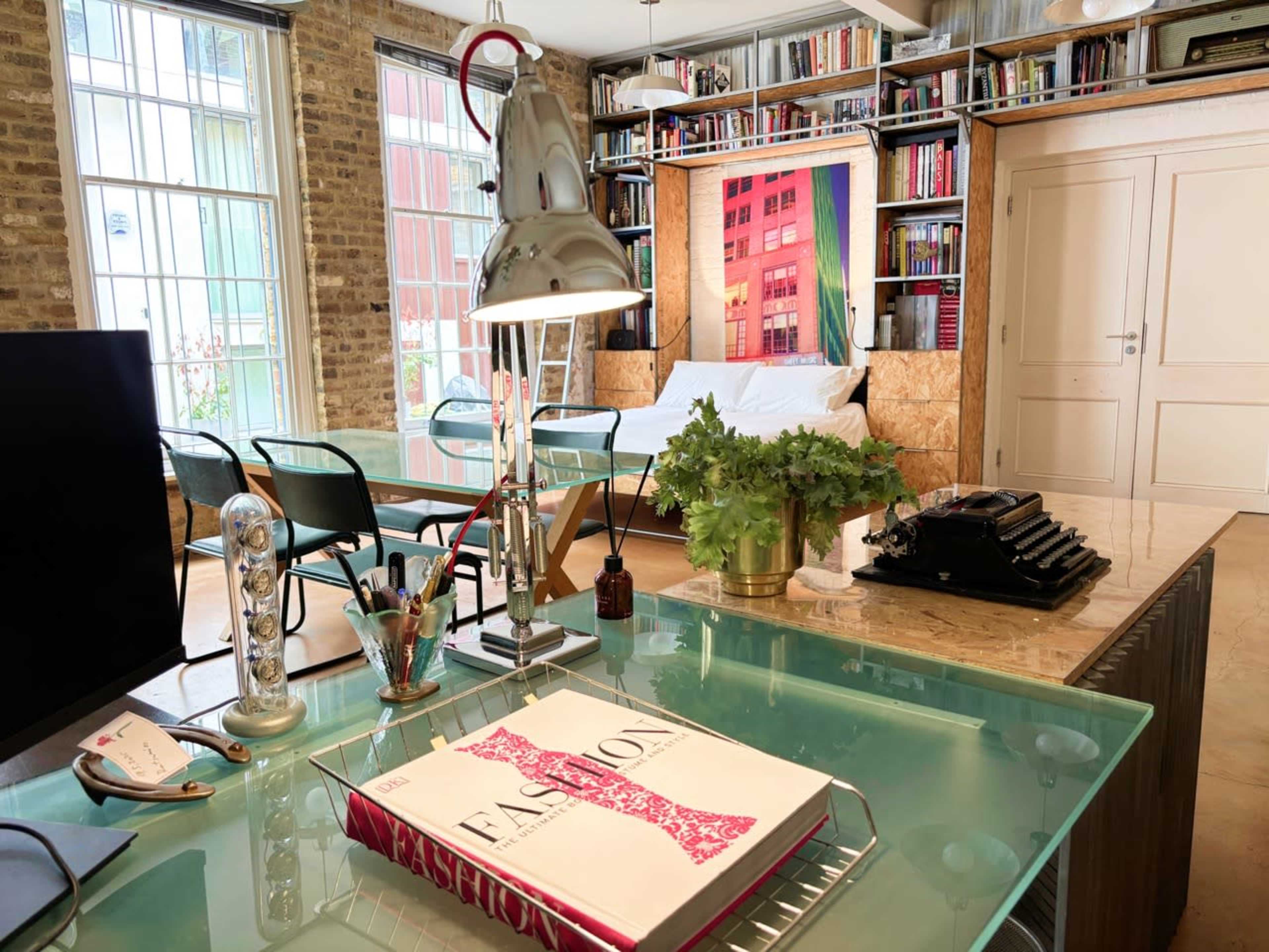 The image shows a bright, modern living space with brick walls, a glass table, a typewriter on a kitchen countertop, and bookshelves filled with various books.