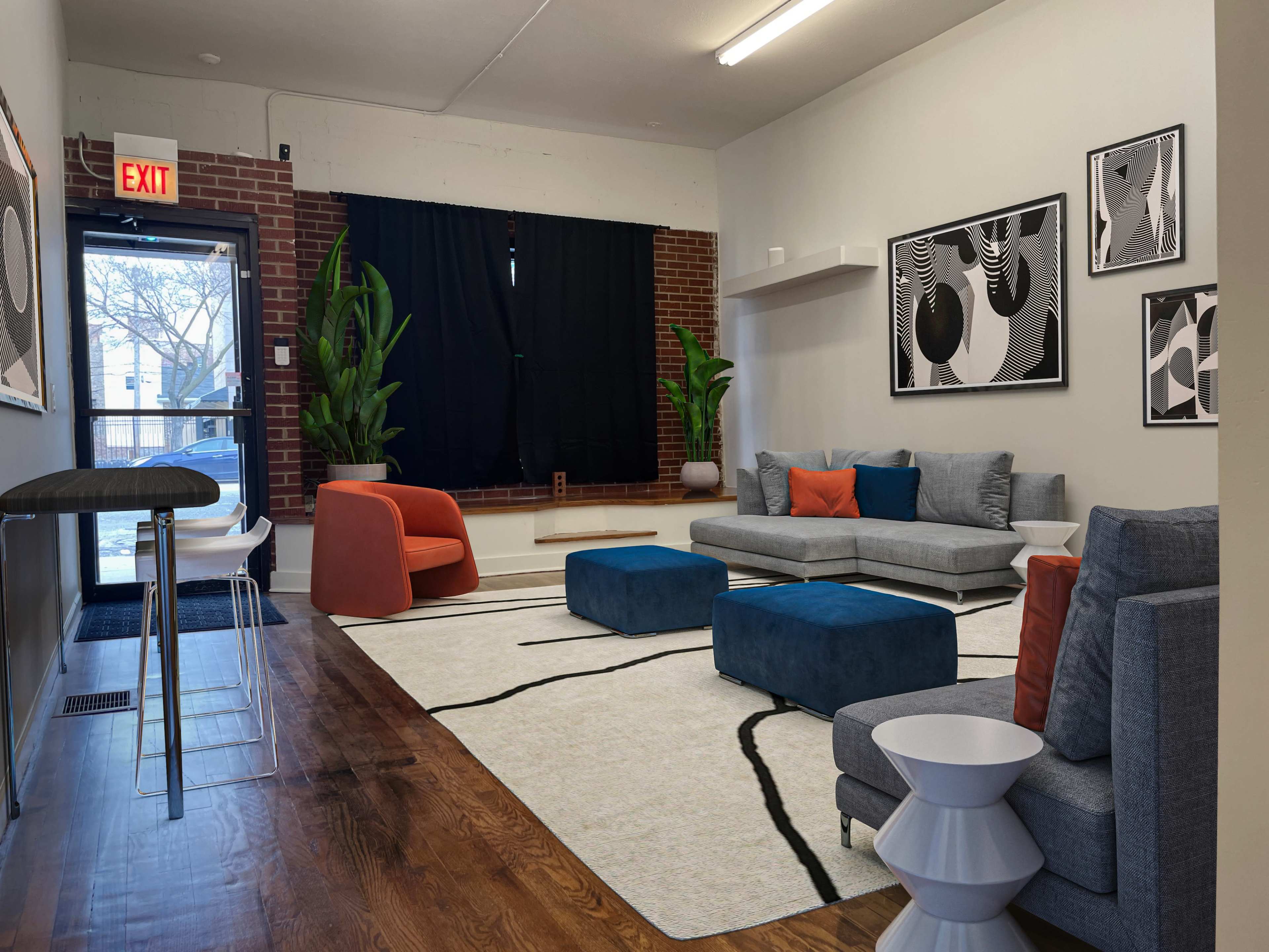 A lightly furnished lounge area with a gray sectional sofa, an orange accent chair, two ottomans, a patterned rug, and plants, all set against a backdrop of exposed brick and minimalist artwork.