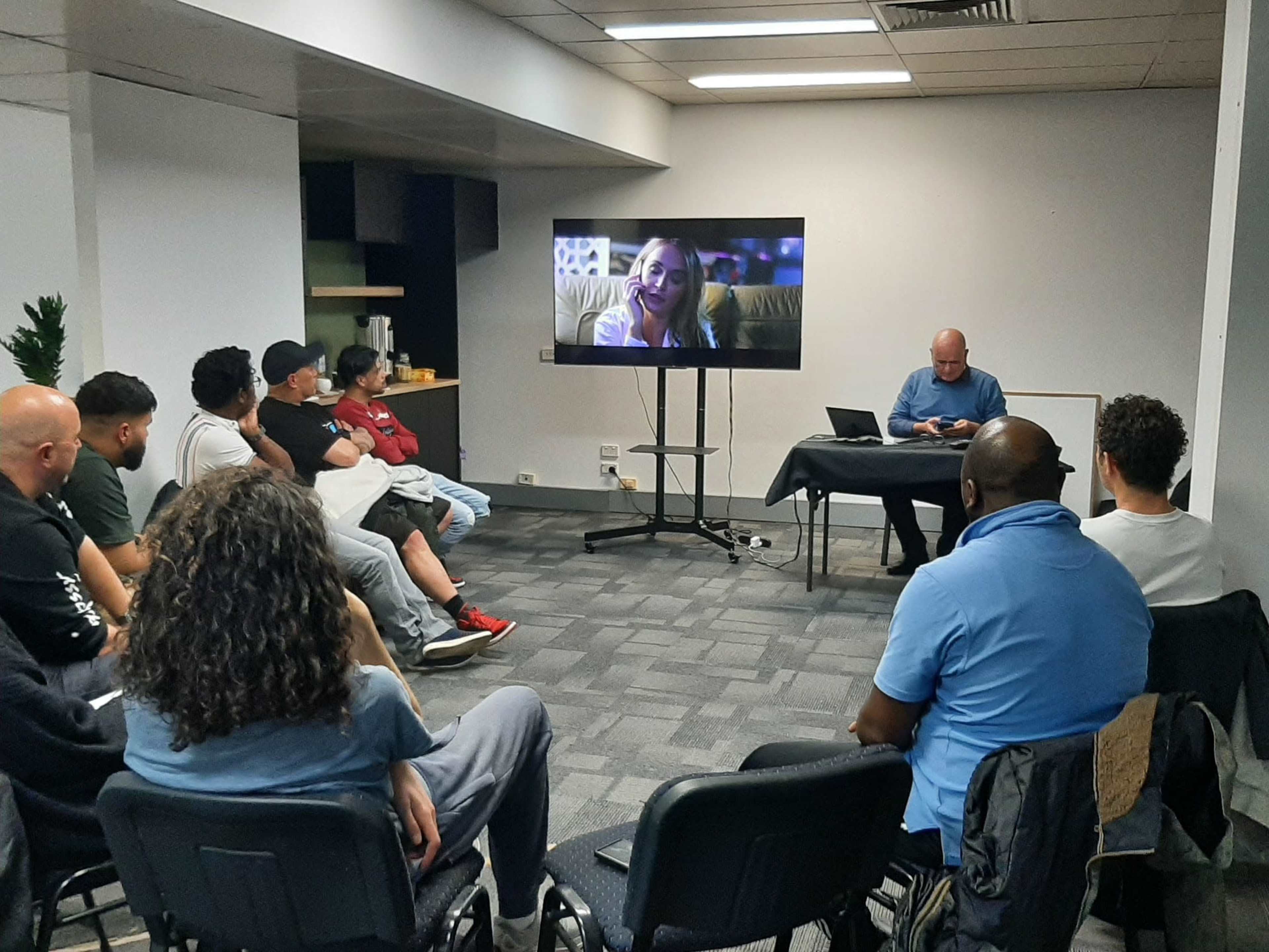 A group of people sits in a room, watching a video on a large screen while another person works at a table nearby.