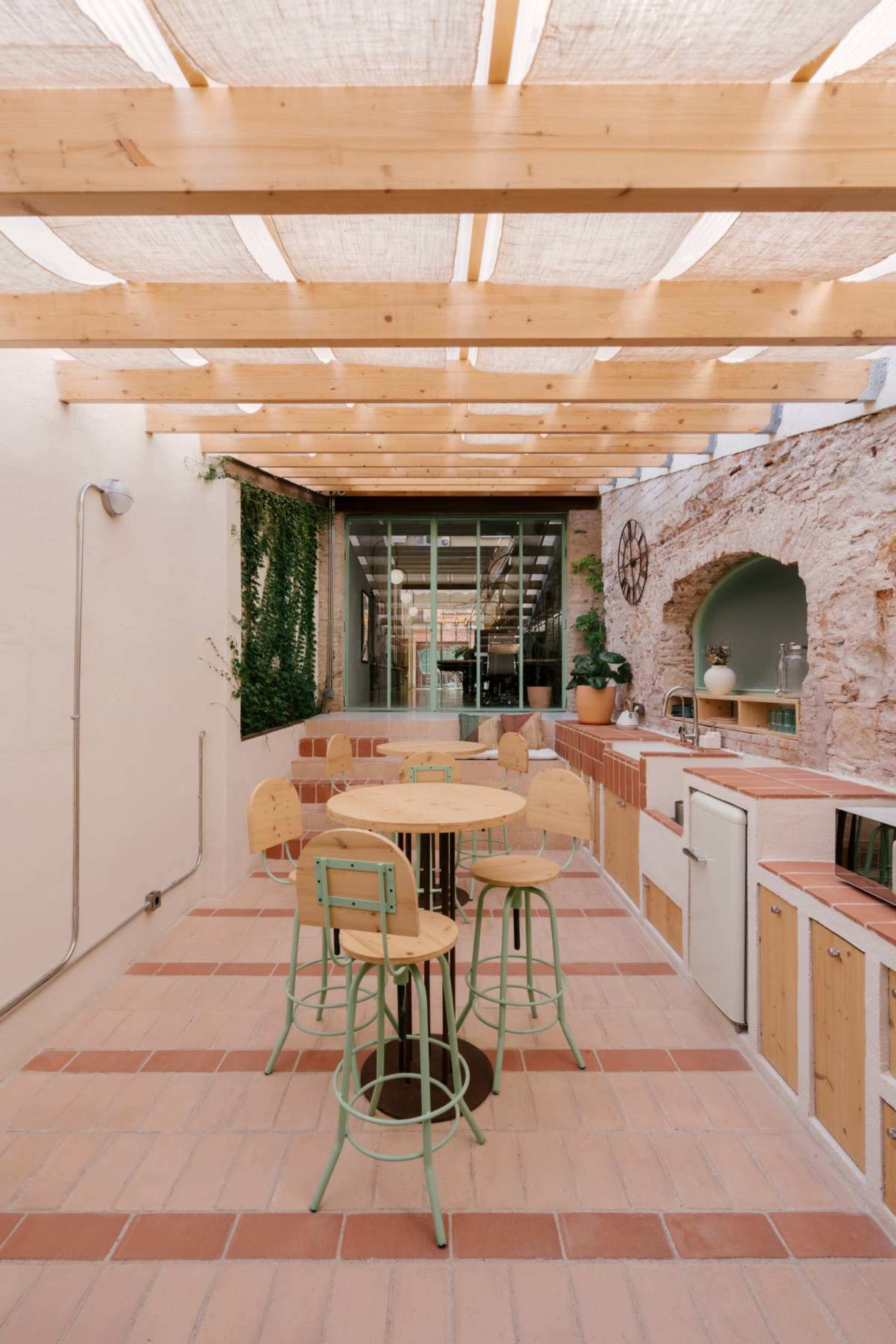 The image shows a modern outdoor kitchen area with wooden beams overhead and a table surrounded by stools.