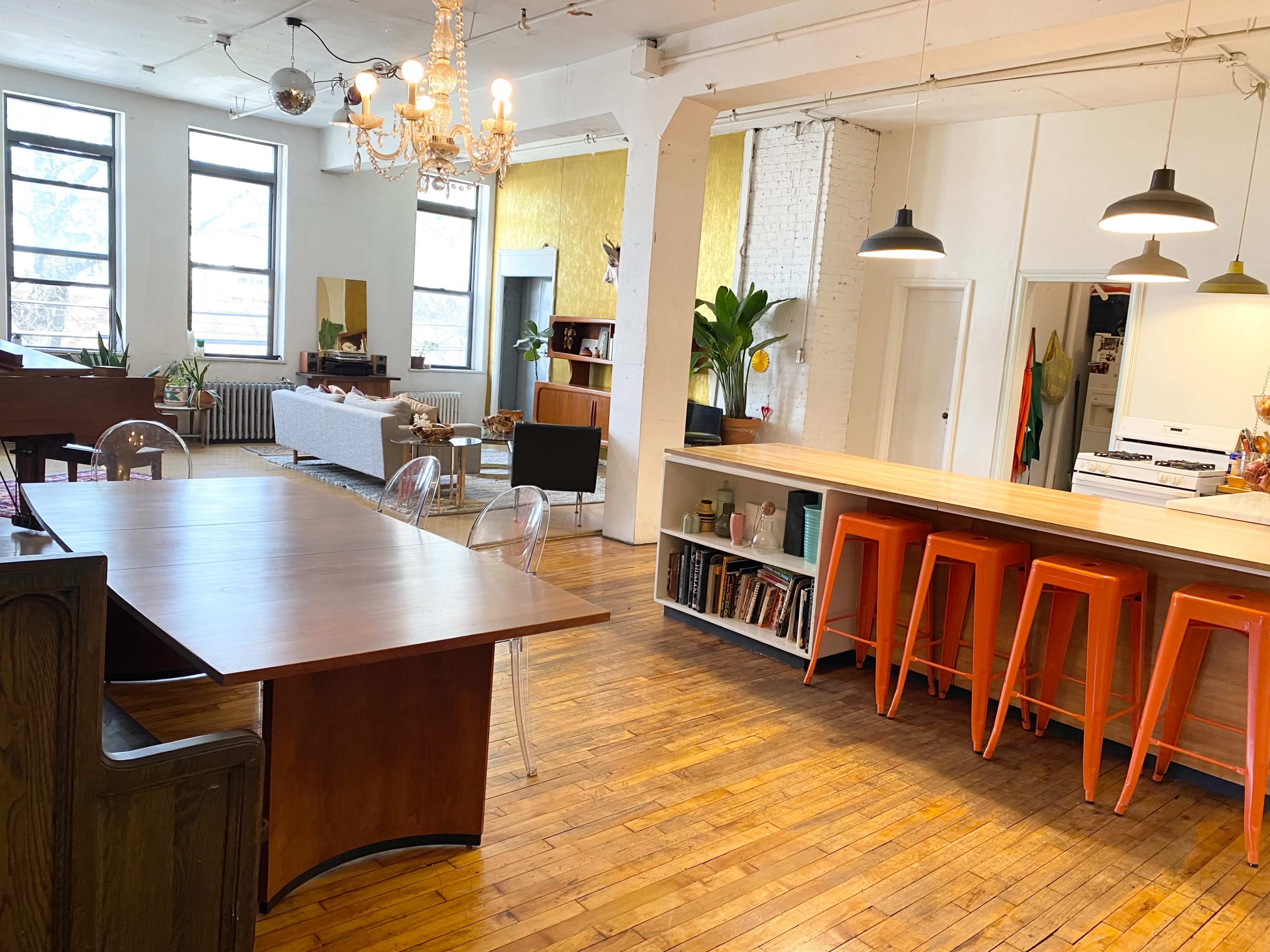 A bright, open living space featuring a wooden dining table, a kitchen with orange stools, and large windows allowing natural light to fill the room.