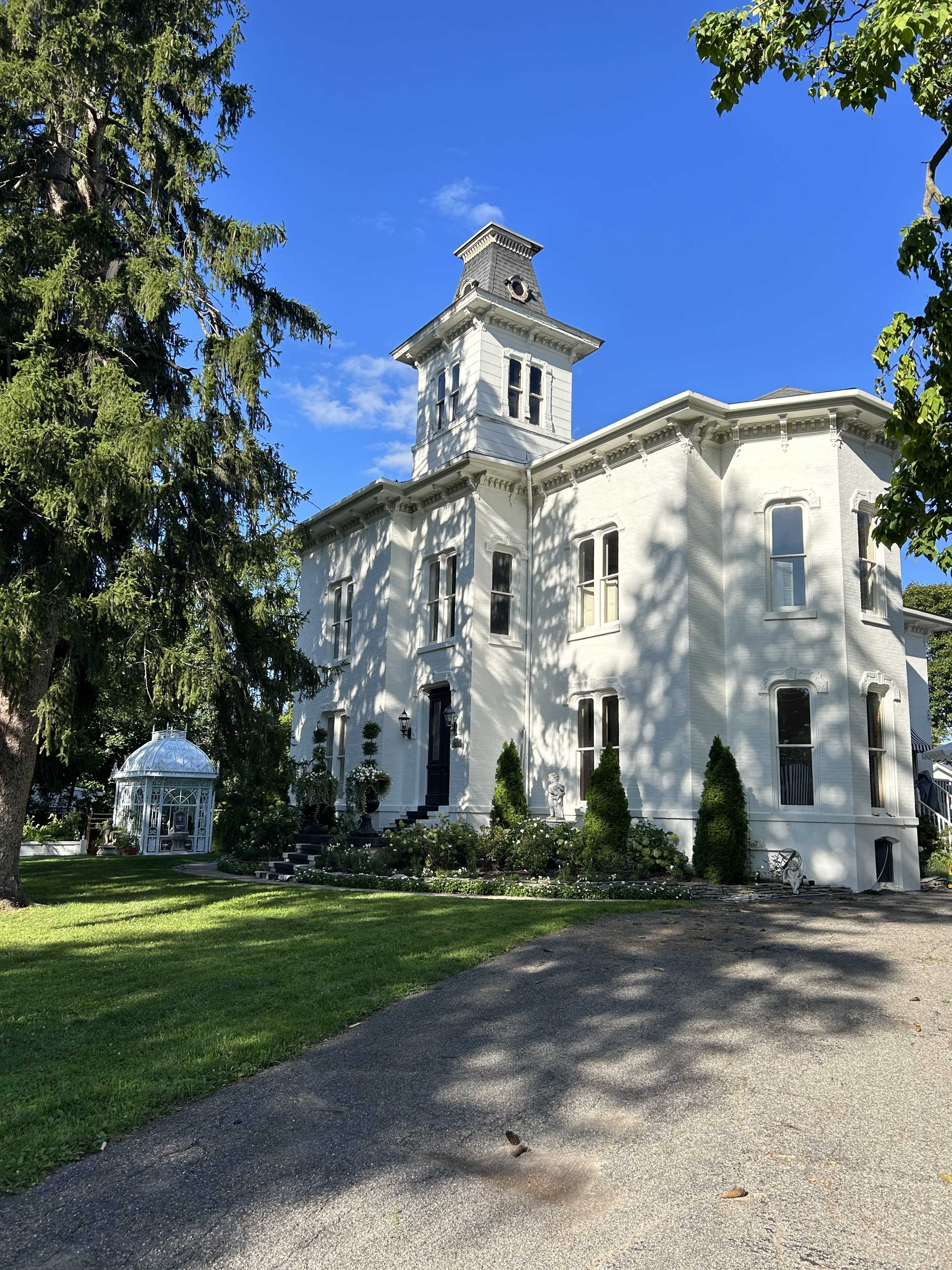 The image shows a large white historic house with a clock tower, surrounded by green grass and trees, and a small glass greenhouse nearby.