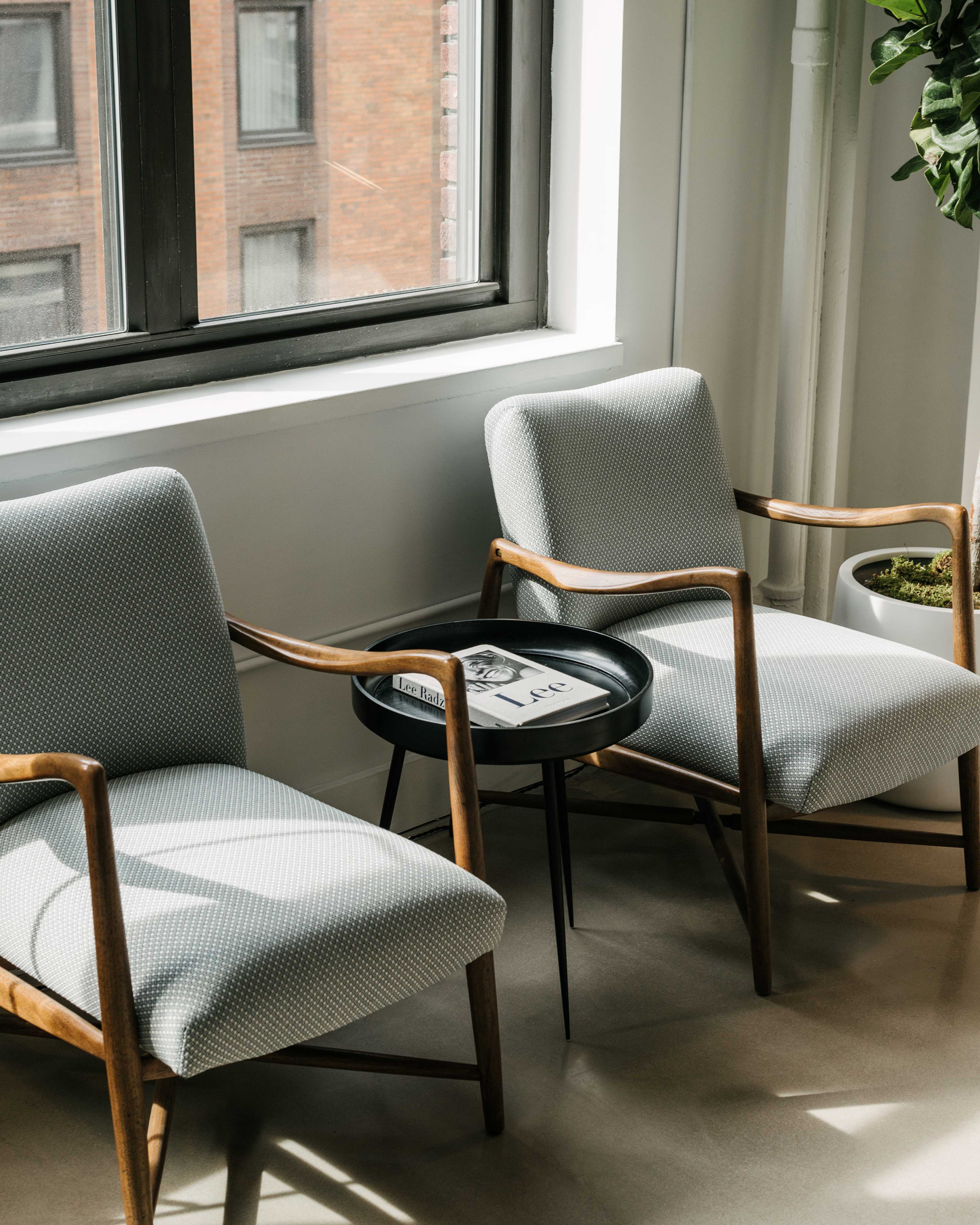 Two mid-century modern chairs with patterned fabric are positioned beside a small round table, next to a large window illuminated by natural light.