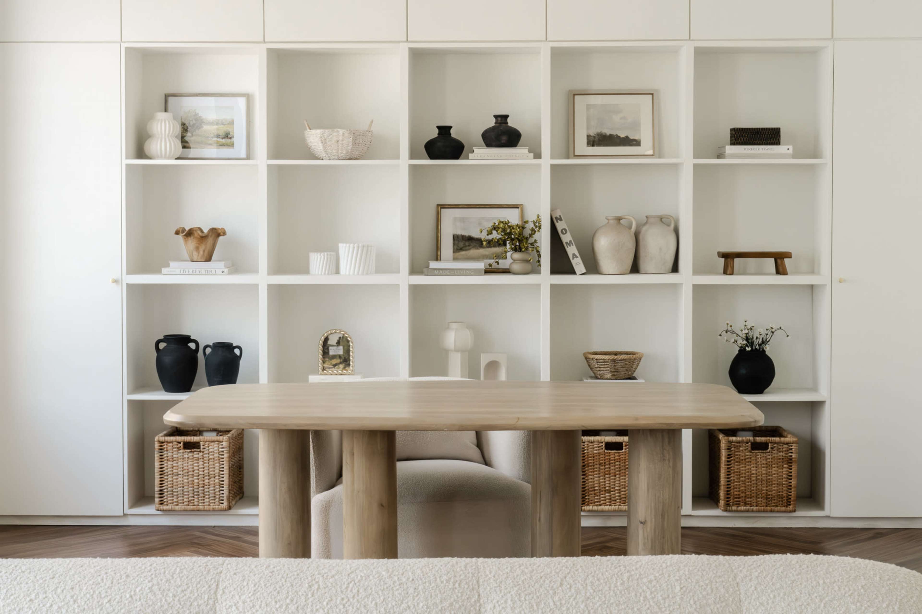 The image features a modern interior with a wooden table in front of a white shelving unit displaying various decorative objects and baskets.