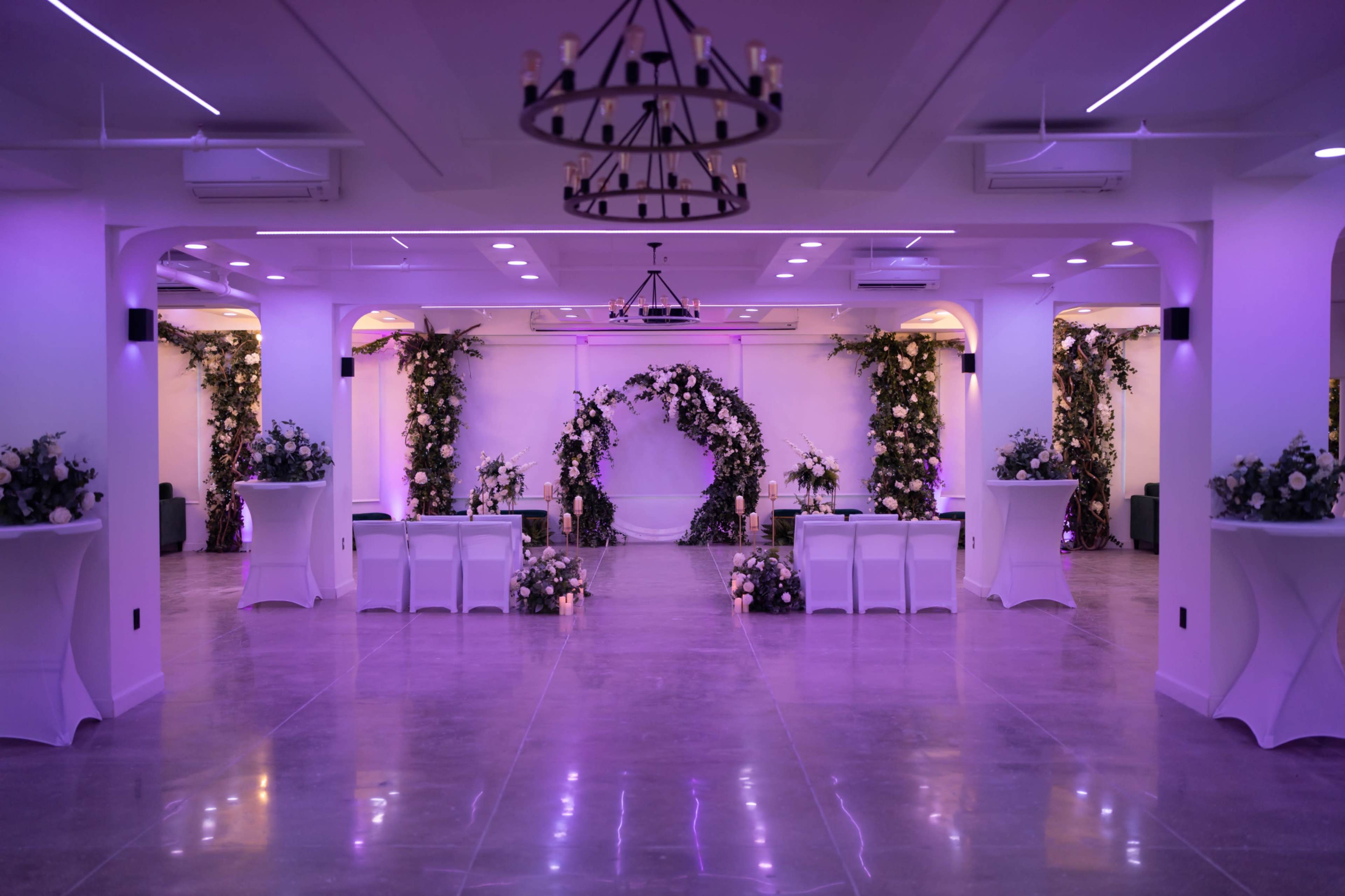 A spacious, elegantly decorated venue features floral arrangements and a circular backdrop, illuminated by purple lighting.
