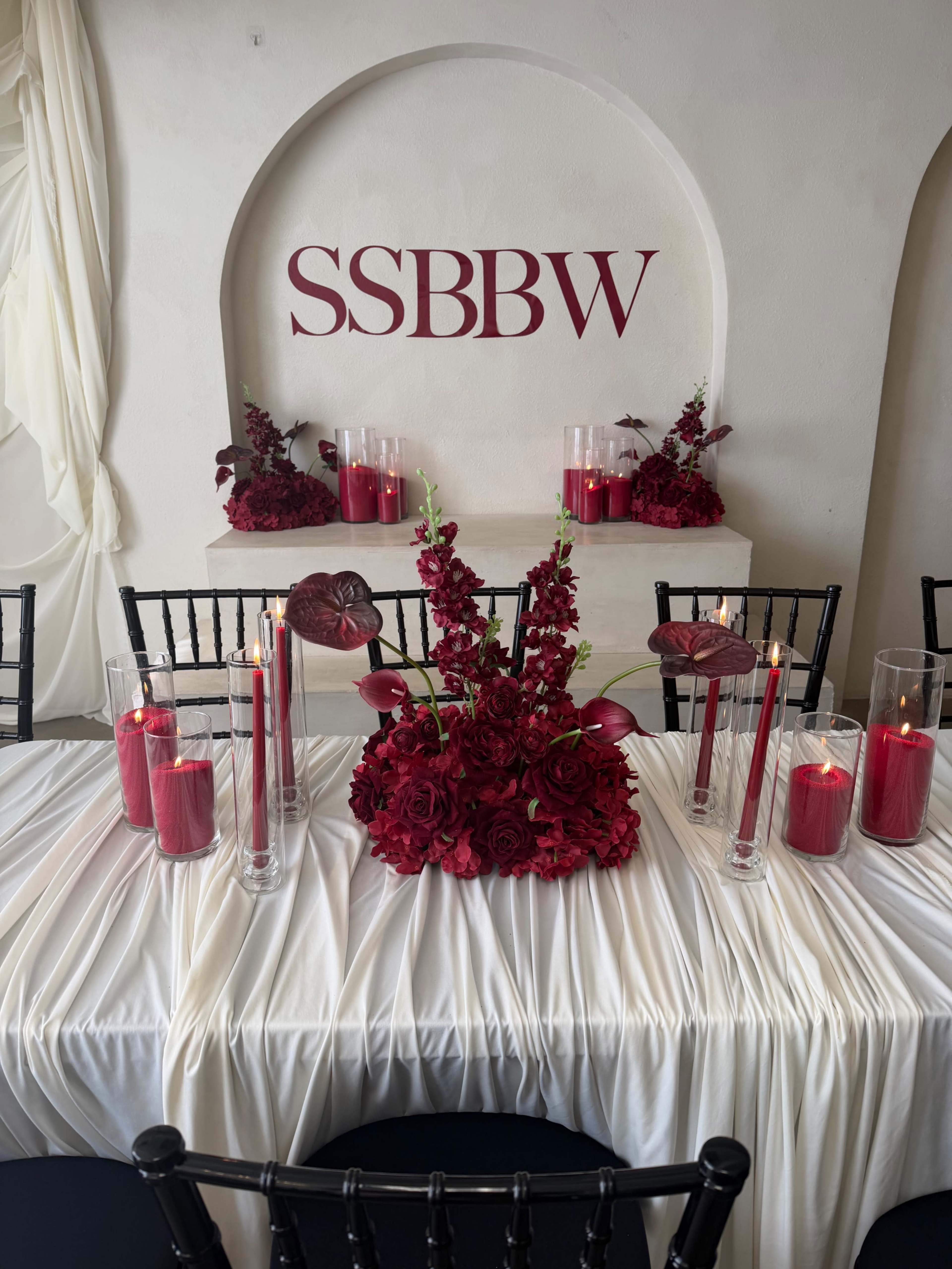 The scene features a elegantly set table draped in white fabric, adorned with a centerpiece of deep red flowers, candles, and a backdrop displaying the initials "SSBBBW."