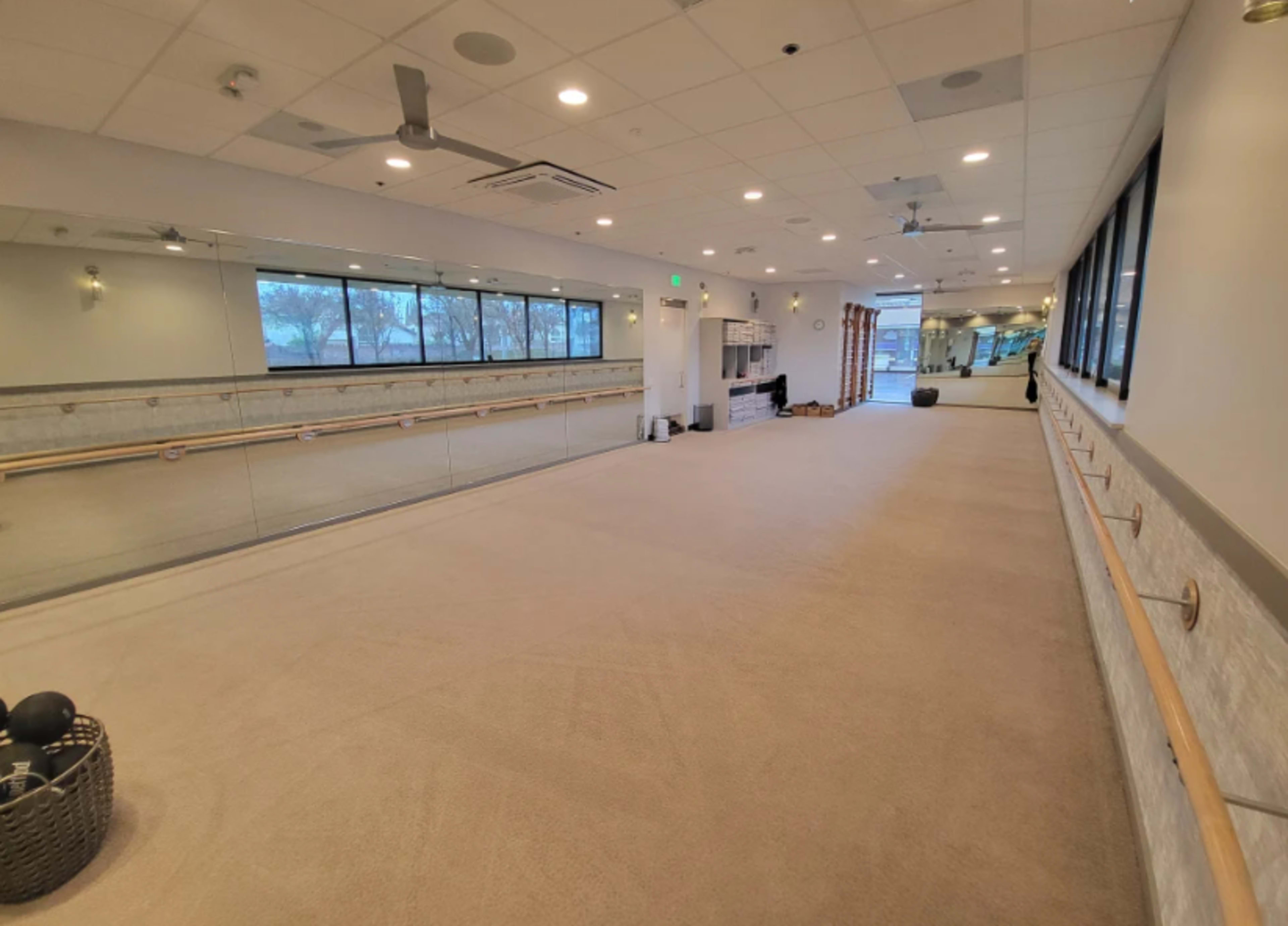 The image shows an empty fitness studio with mirrors along one wall and a barre installed across the room.