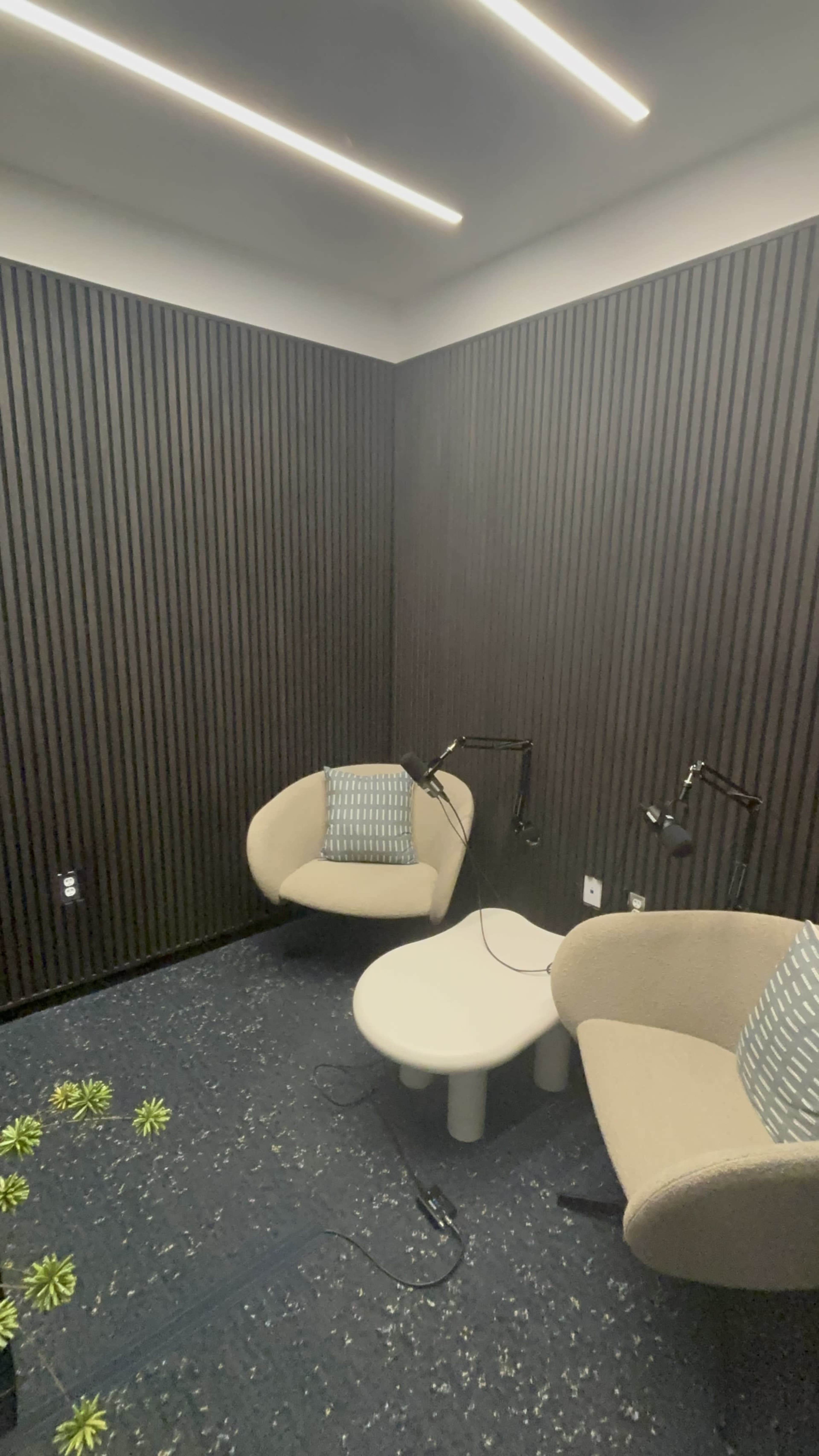 The image shows a small, modern meeting room with two beige chairs, a round white table, and wall-mounted microphones, surrounded by dark wood paneling.