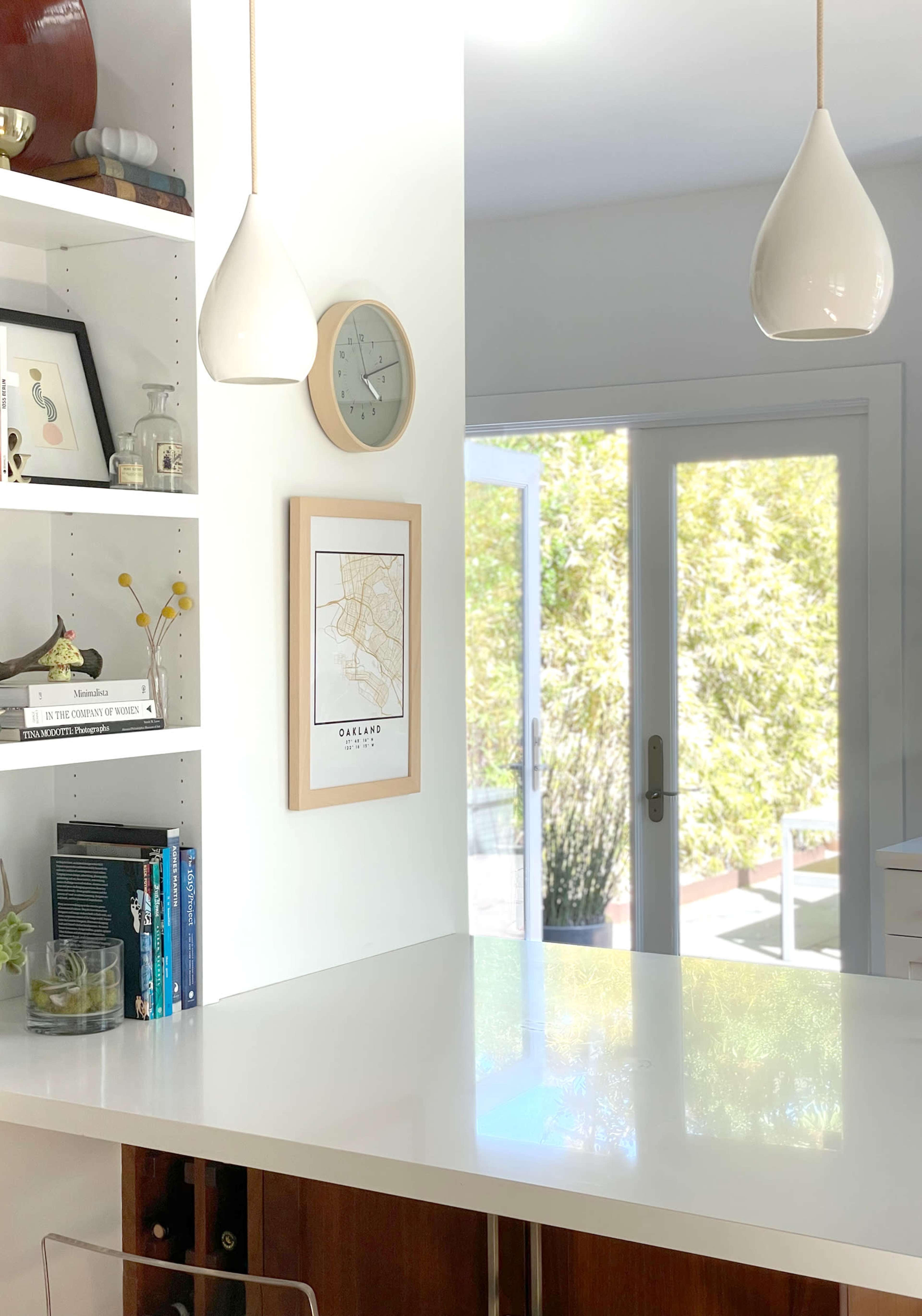 A modern kitchen area with a white countertop, decorative shelves, a wall clock, and a framed map, leading to an outdoor space through glass doors.