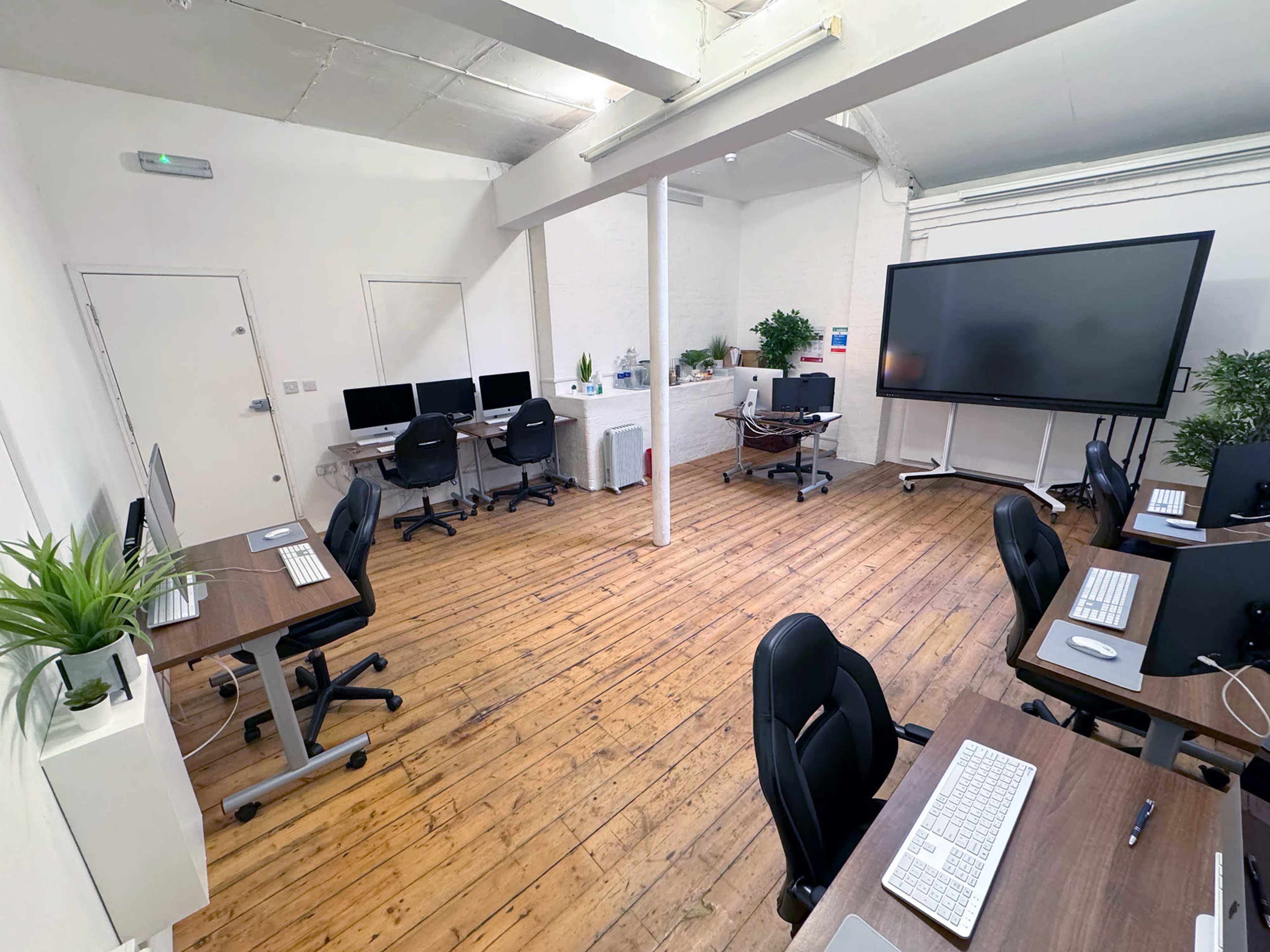 A spacious office with several workstations, computers, and a large screen, surrounded by plants and wooden flooring.