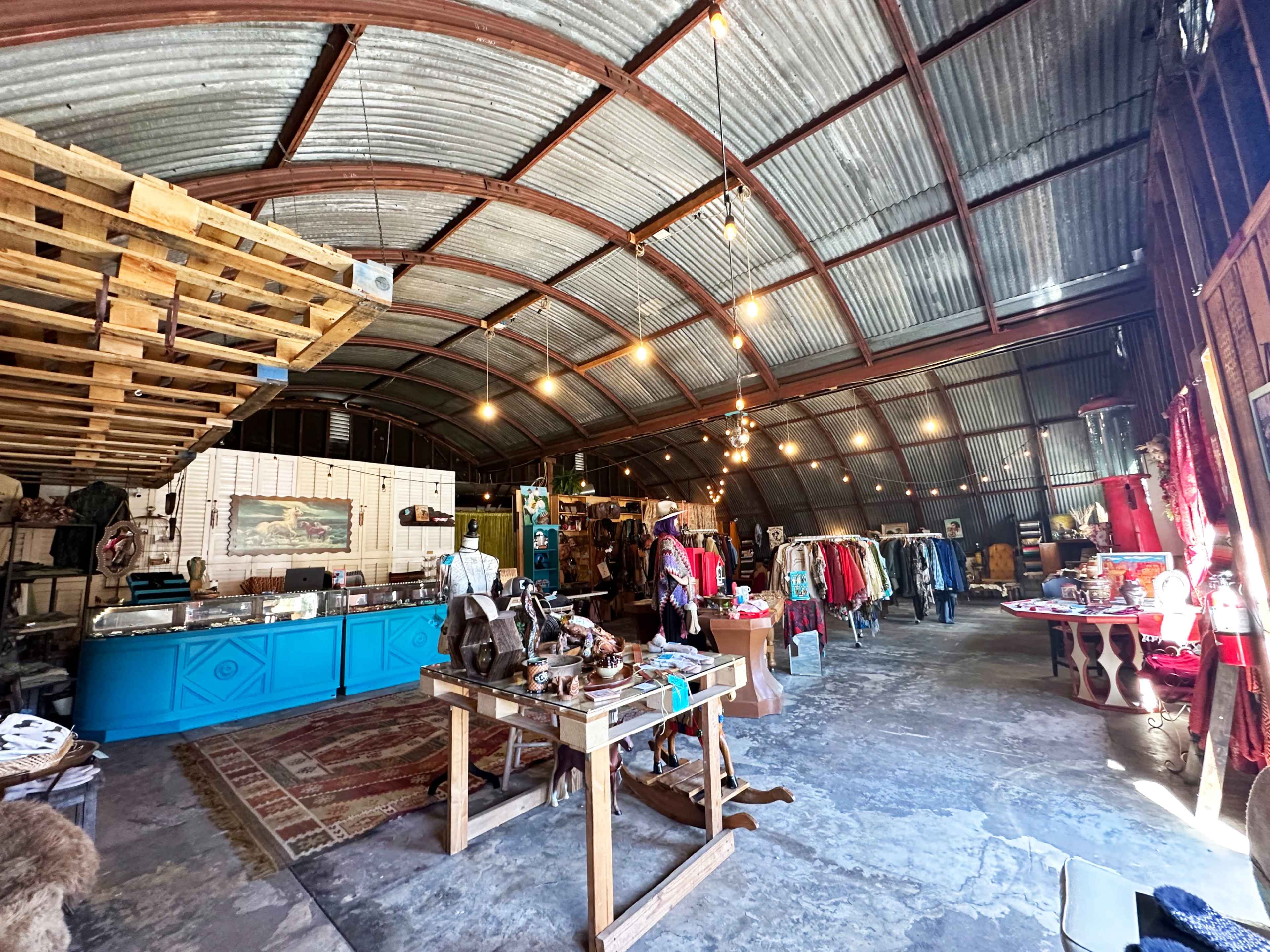 The interior of a rustic warehouse features various items for sale, including clothing on display and a turquoise counter.