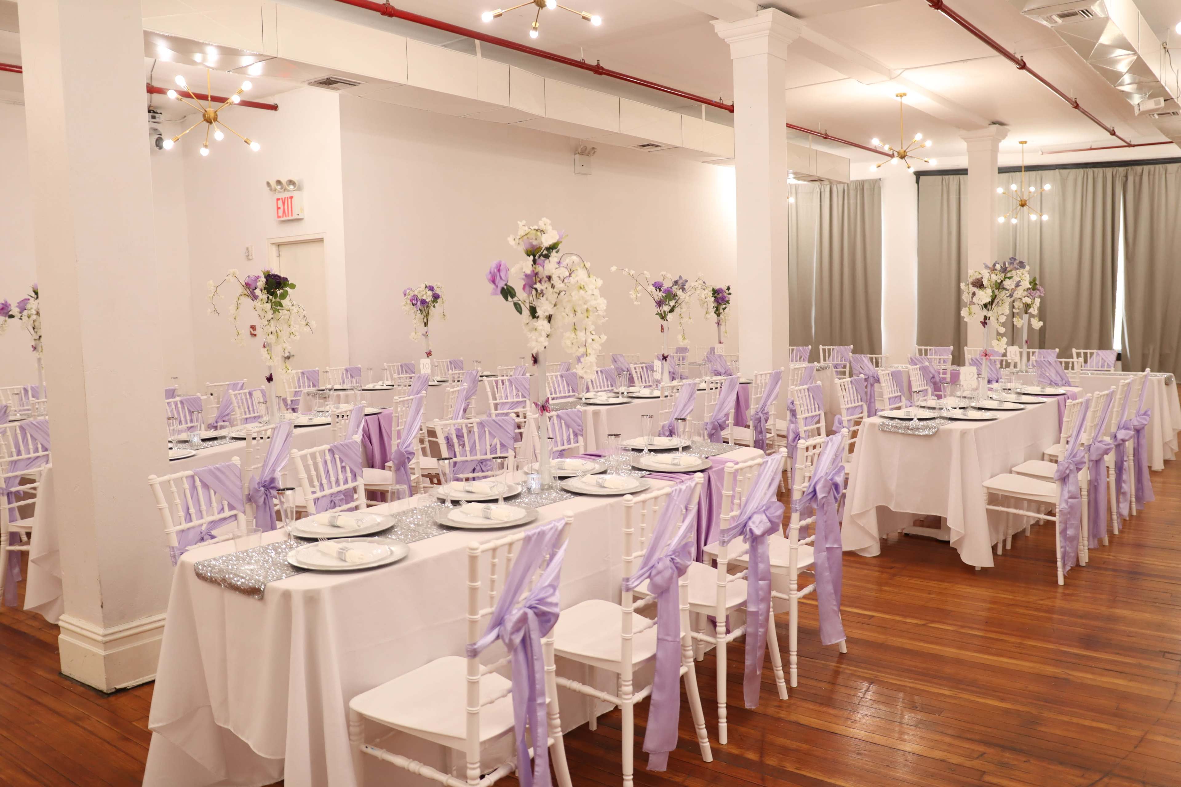An elegantly arranged banquet hall with tables set for an event, featuring white tablecloths, lavender accents, and floral centerpieces.