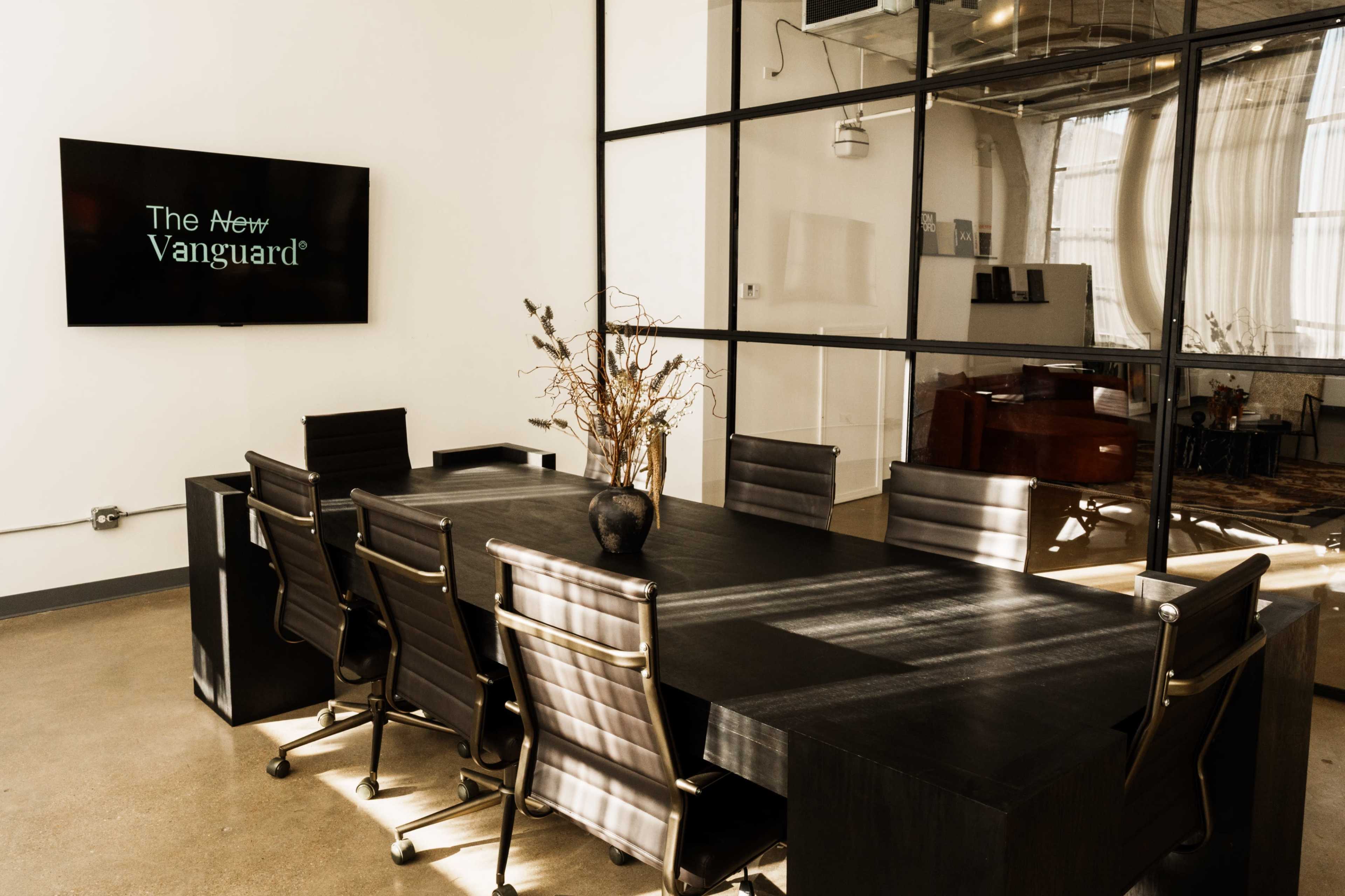 A modern conference room features a long black table surrounded by six chairs, with a large television mounted on the wall displaying the text "The New Vanguard" and large windows providing natural light.