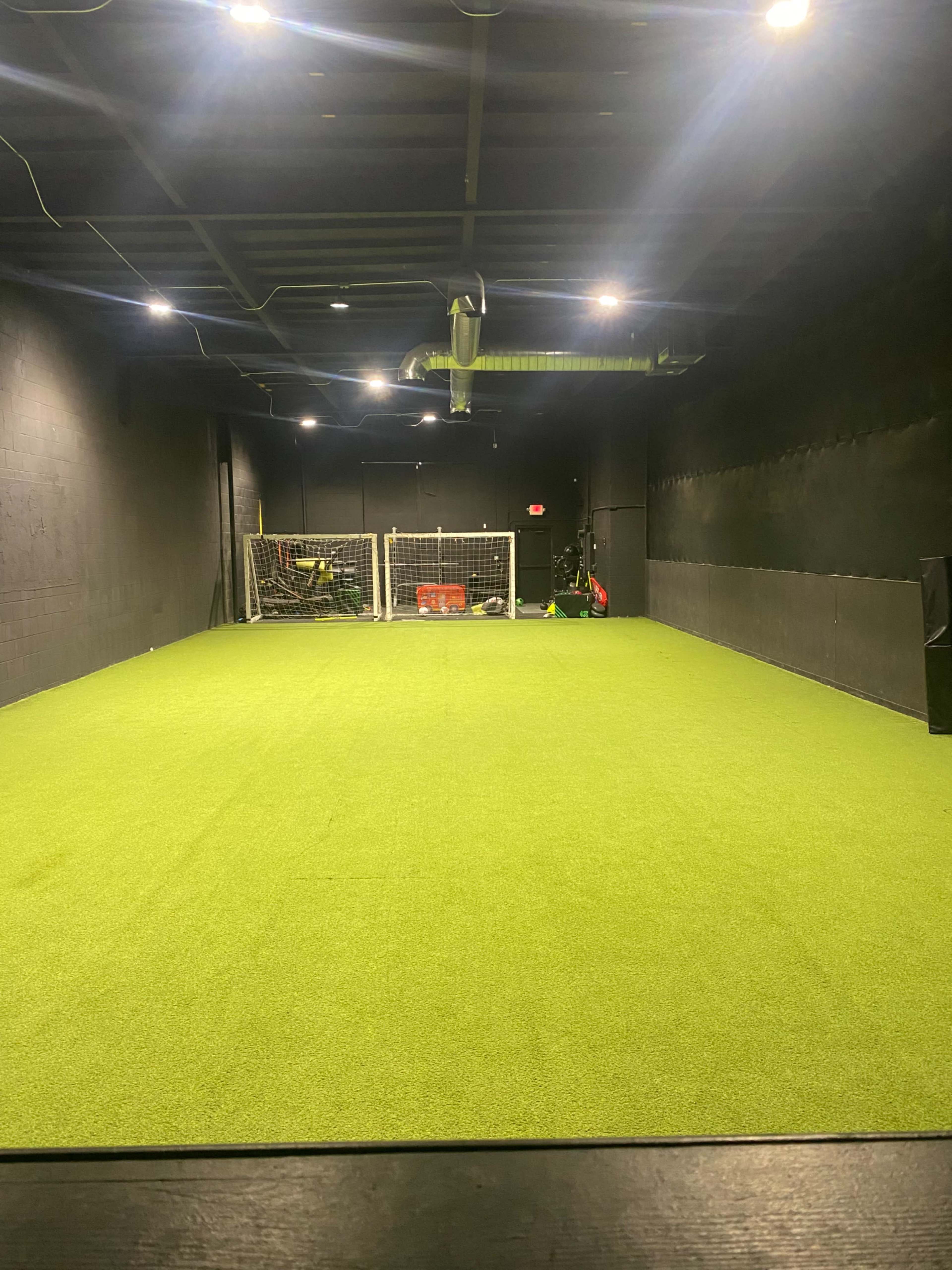 The image shows a spacious indoor training facility with artificial turf, two soccer goals in the background, and track lighting overhead.