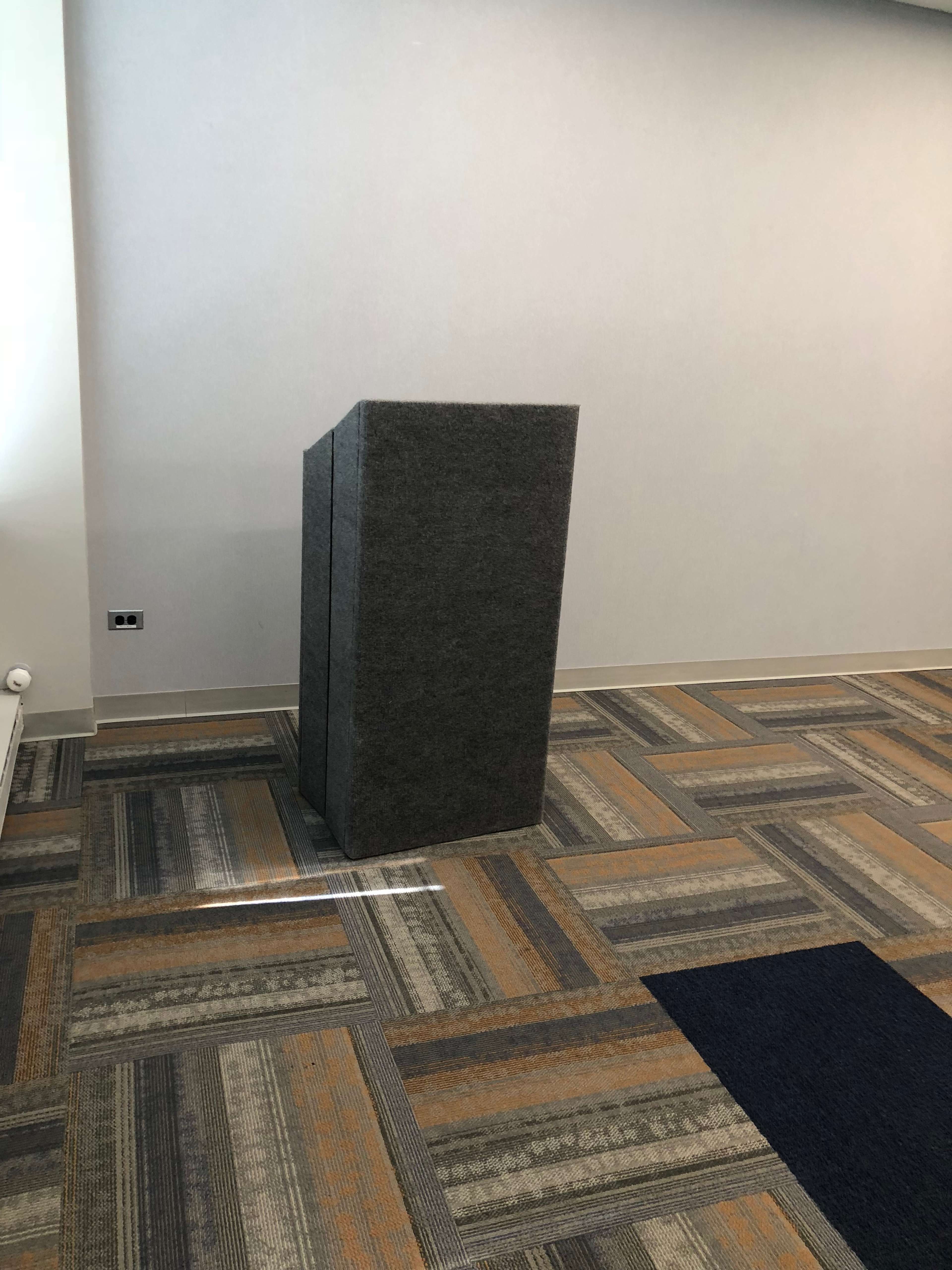 A gray podium stands against a plain wall on a carpeted floor with a rectangular pattern.
