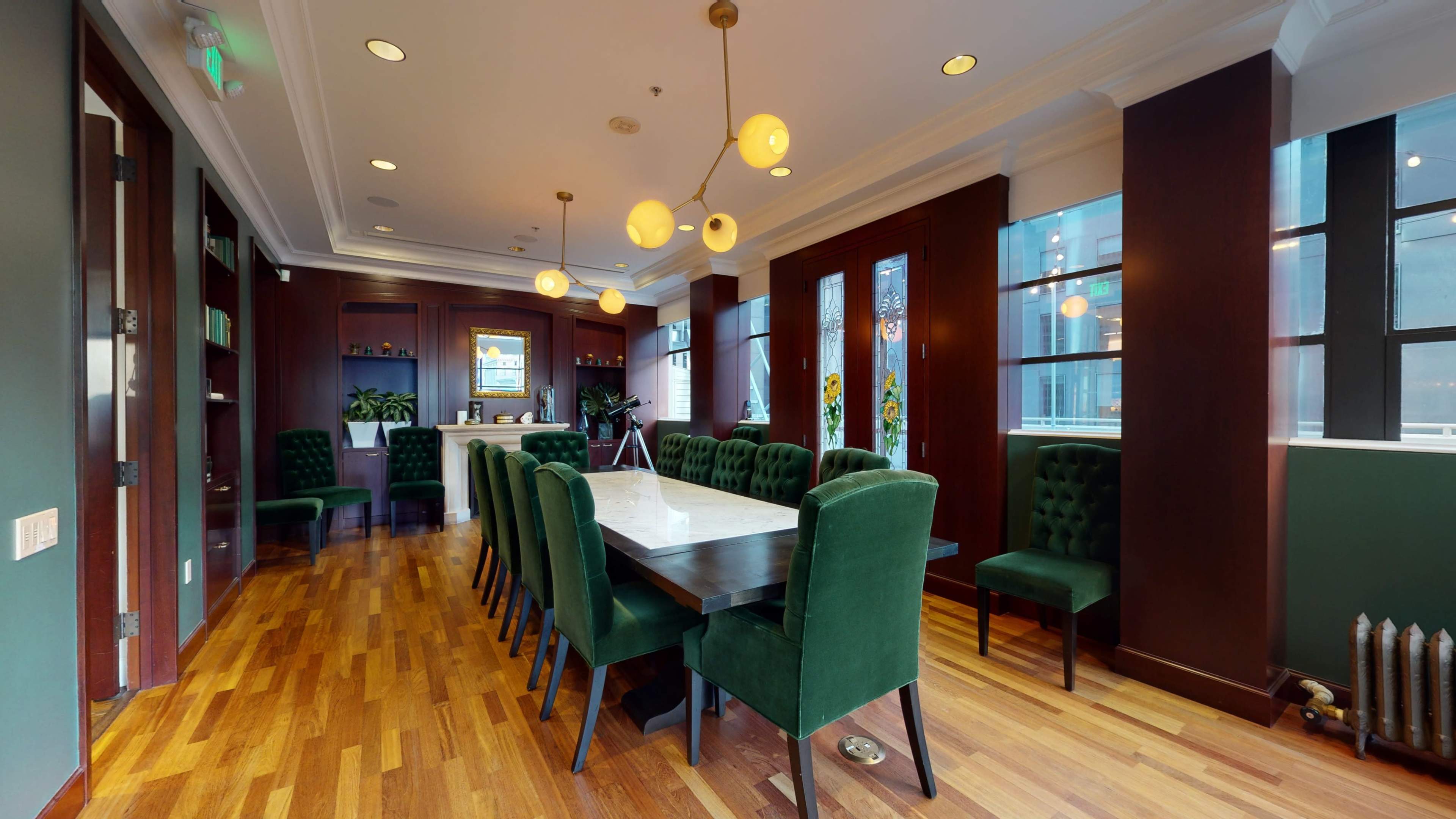 A conference room with a long wooden table surrounded by green upholstered chairs, large windows, and decorative walls.