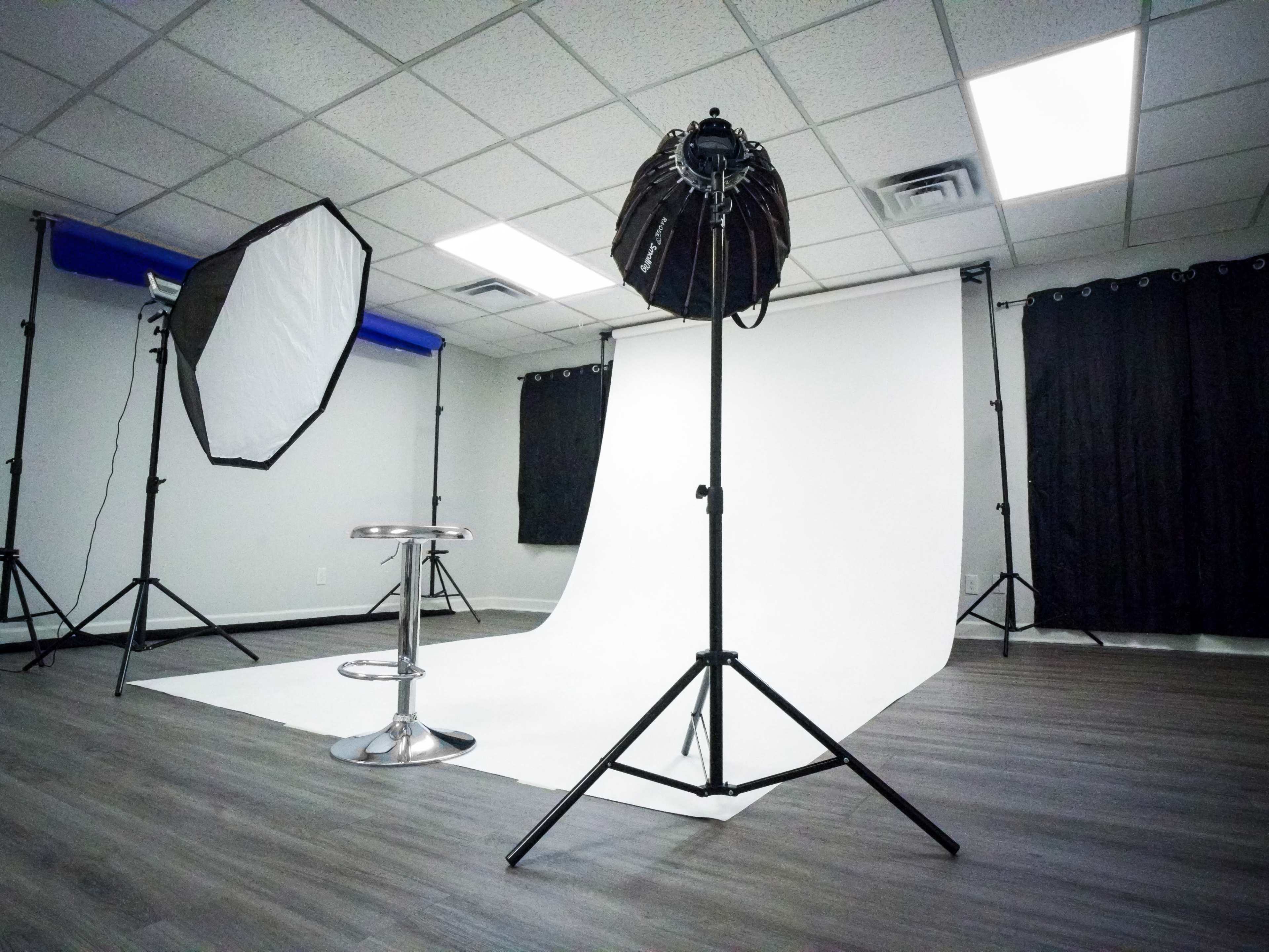 The image shows a photography studio with a white backdrop, softbox lights, a silver stool, and dark curtains.