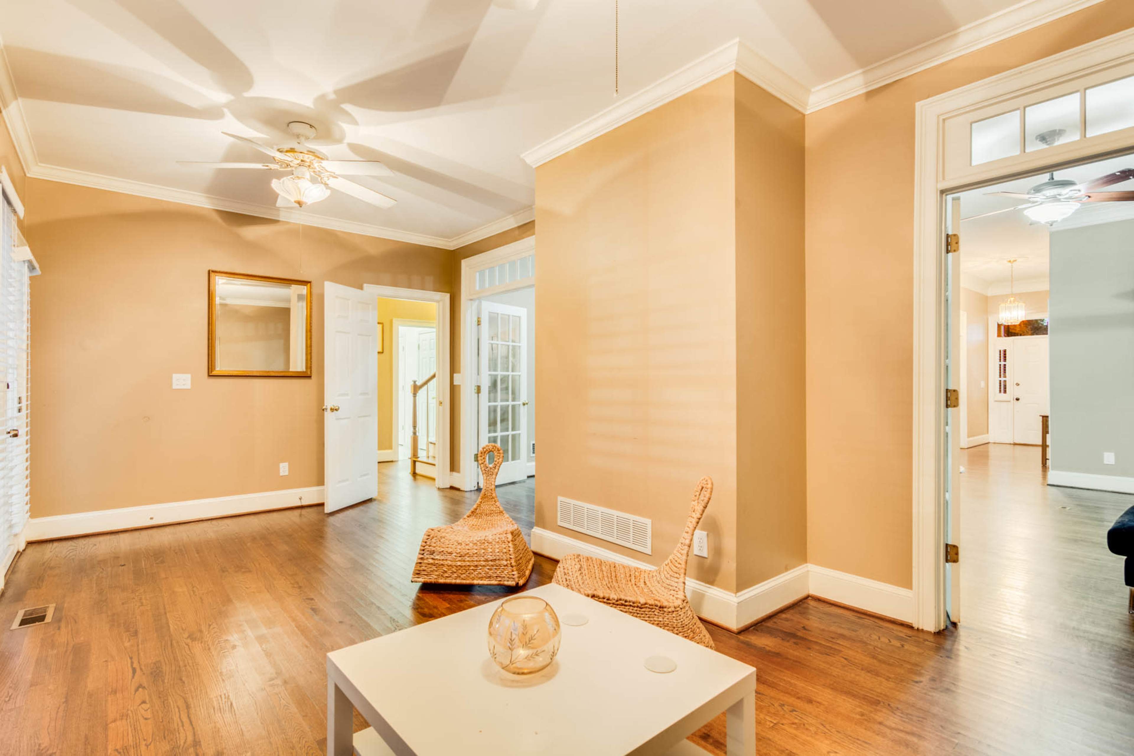The image shows a well-lit living space with a light-colored wall, wooden flooring, and two wicker chairs positioned around a small white table.