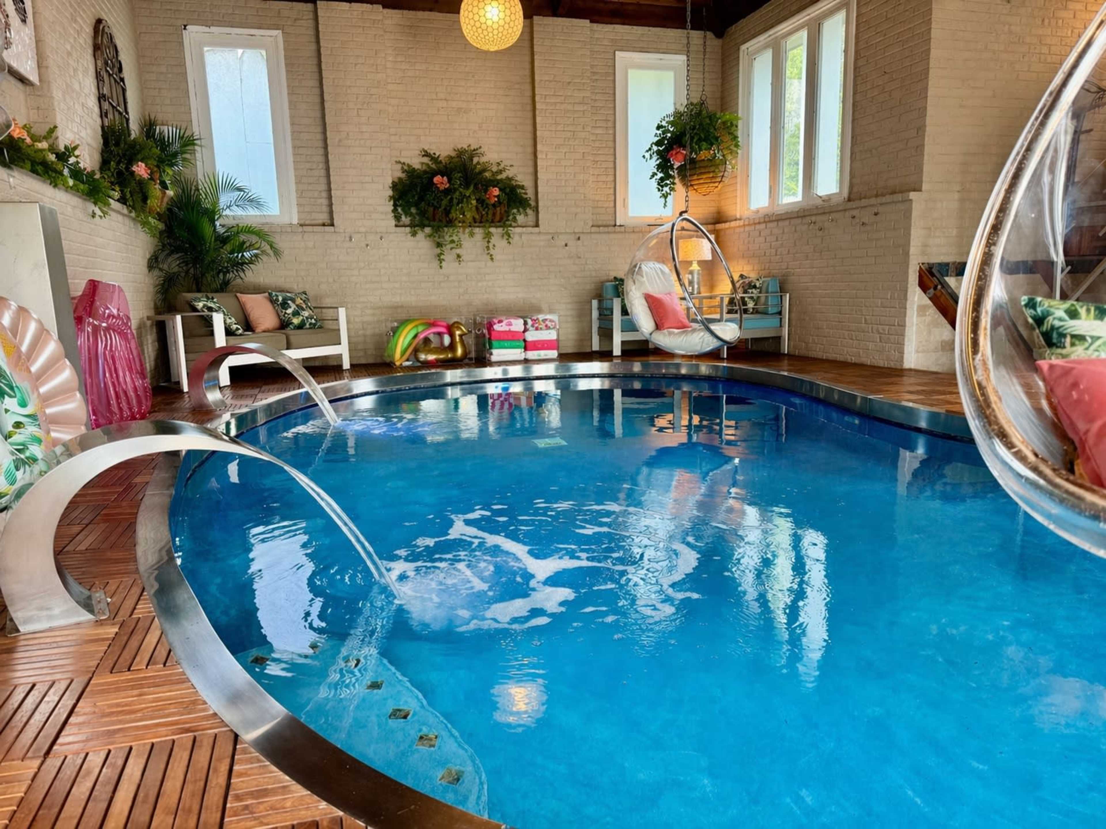 The image shows an indoor pool area with a blue pool, wooden flooring, and modern seating arranged around it.
