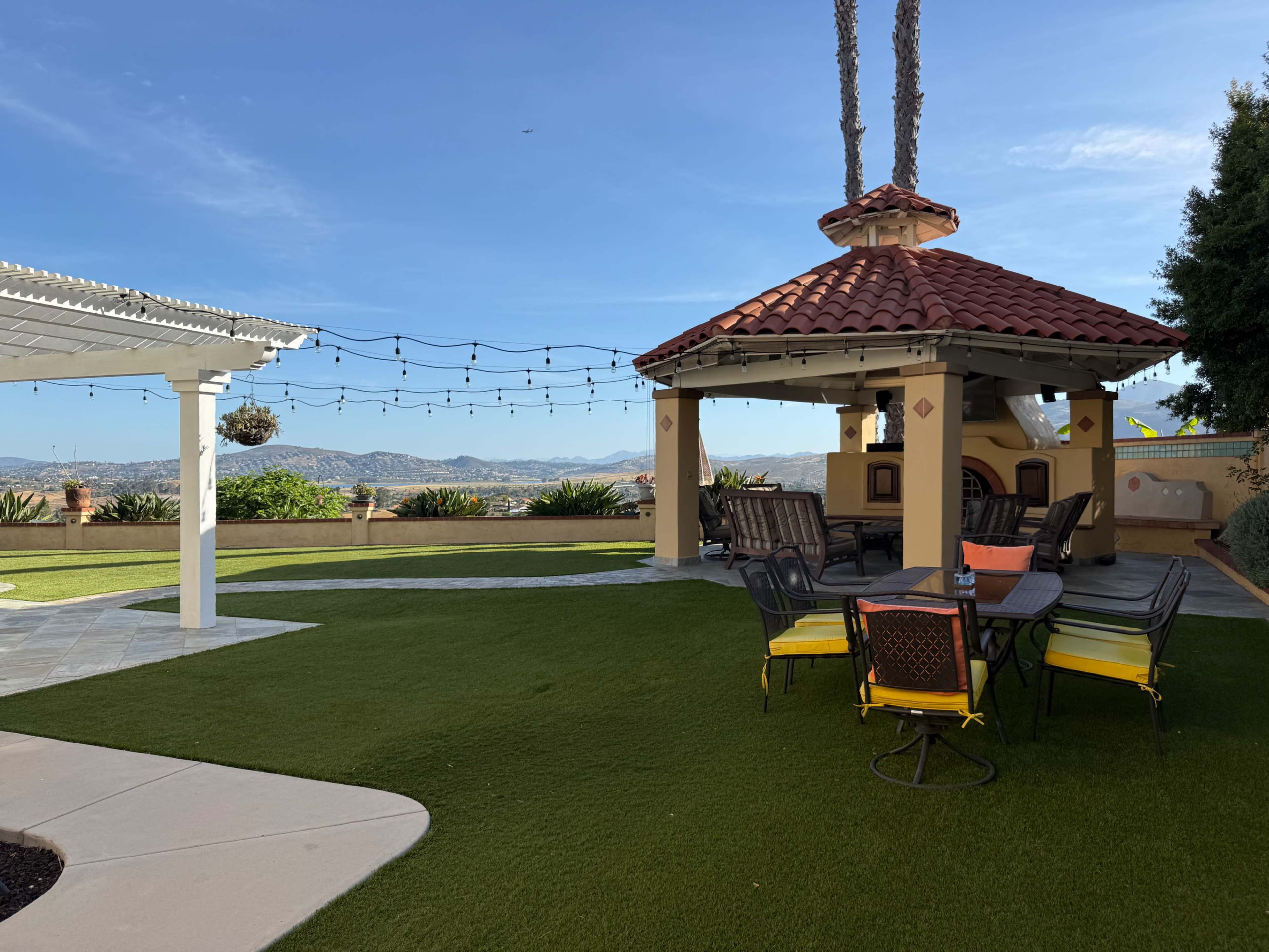The image shows a landscaped backyard featuring a gazebo with a red tile roof, outdoor seating around a table, and a view of distant mountains under a clear blue sky.