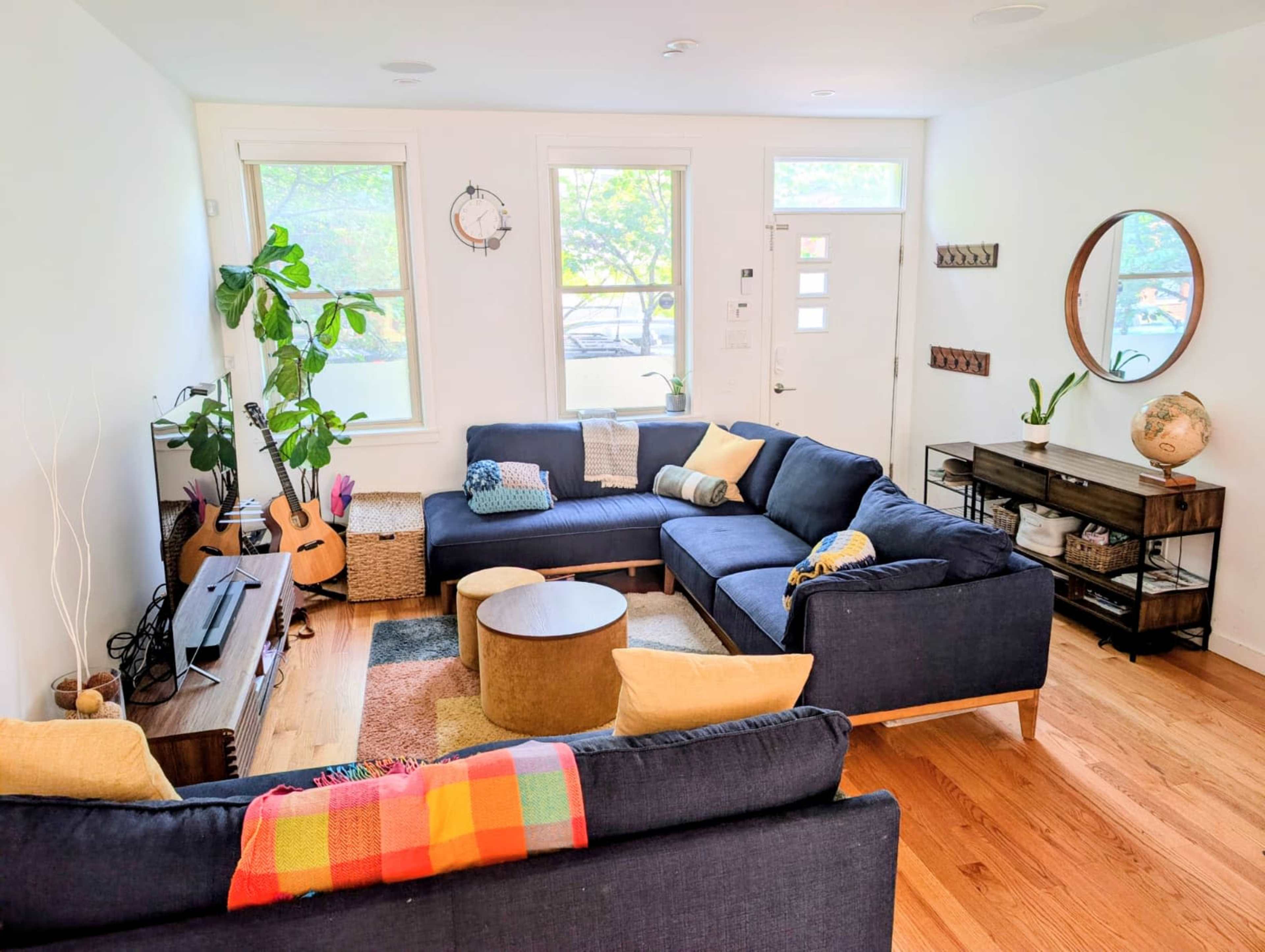 The living room features a large blue sectional sofa, a wooden coffee table, a TV stand, and several plants beside a set of windows.