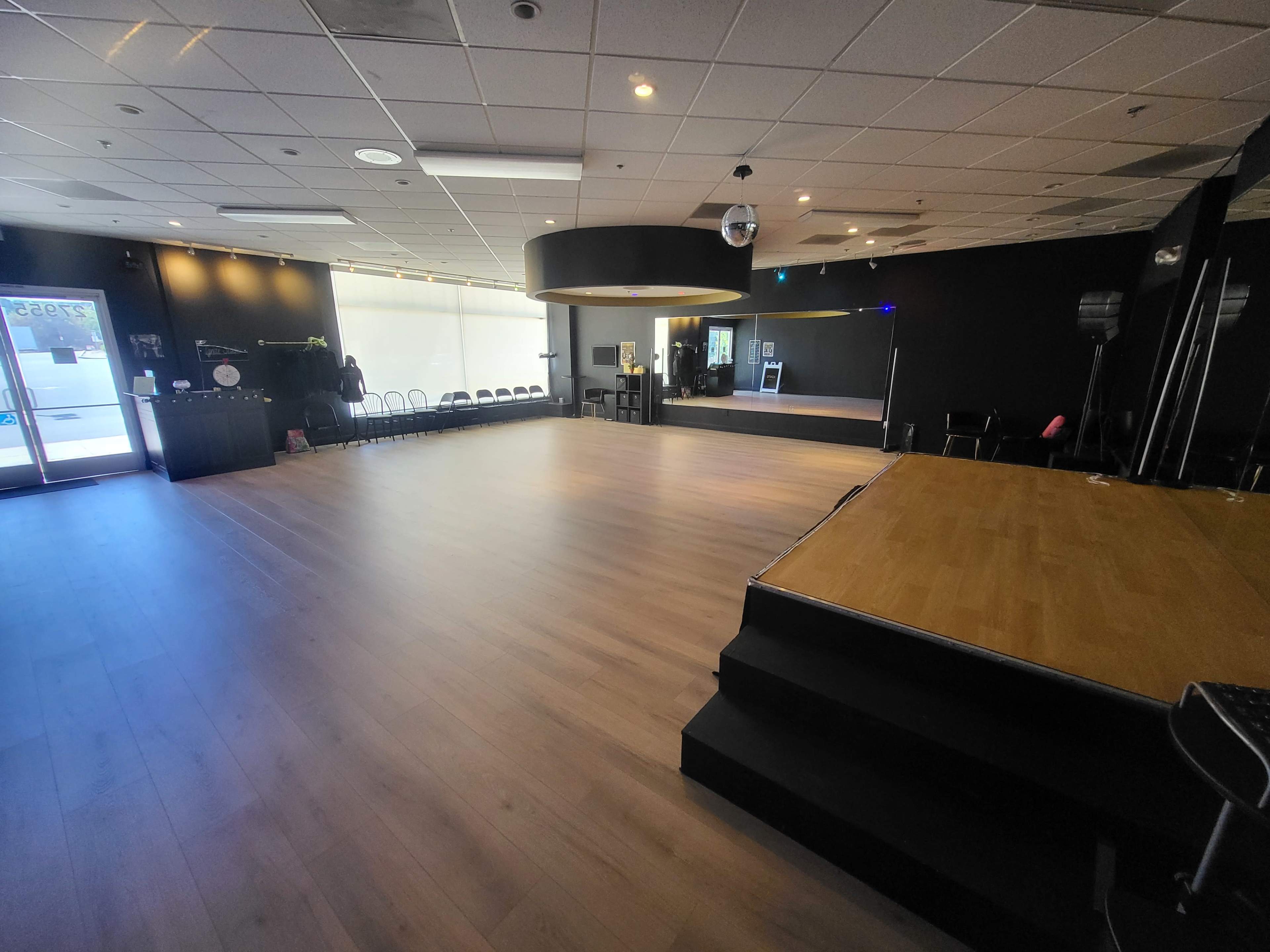 The image shows a spacious, empty dance studio with wooden flooring and a raised stage area in the foreground.