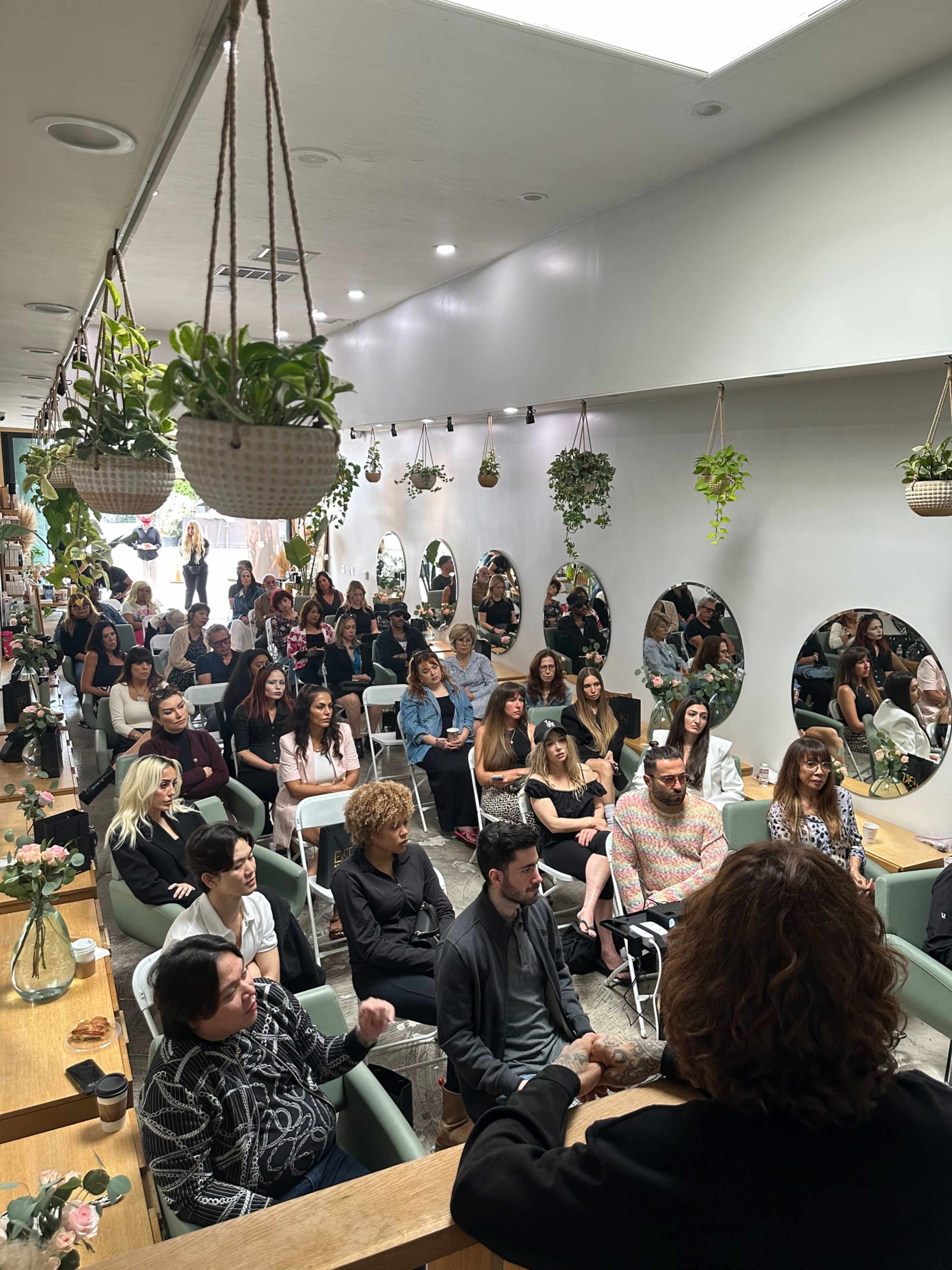 A crowd of attendees is seated in a stylish, plant-filled venue, with large mirrors reflecting their faces as a speaker addresses them from the front.
