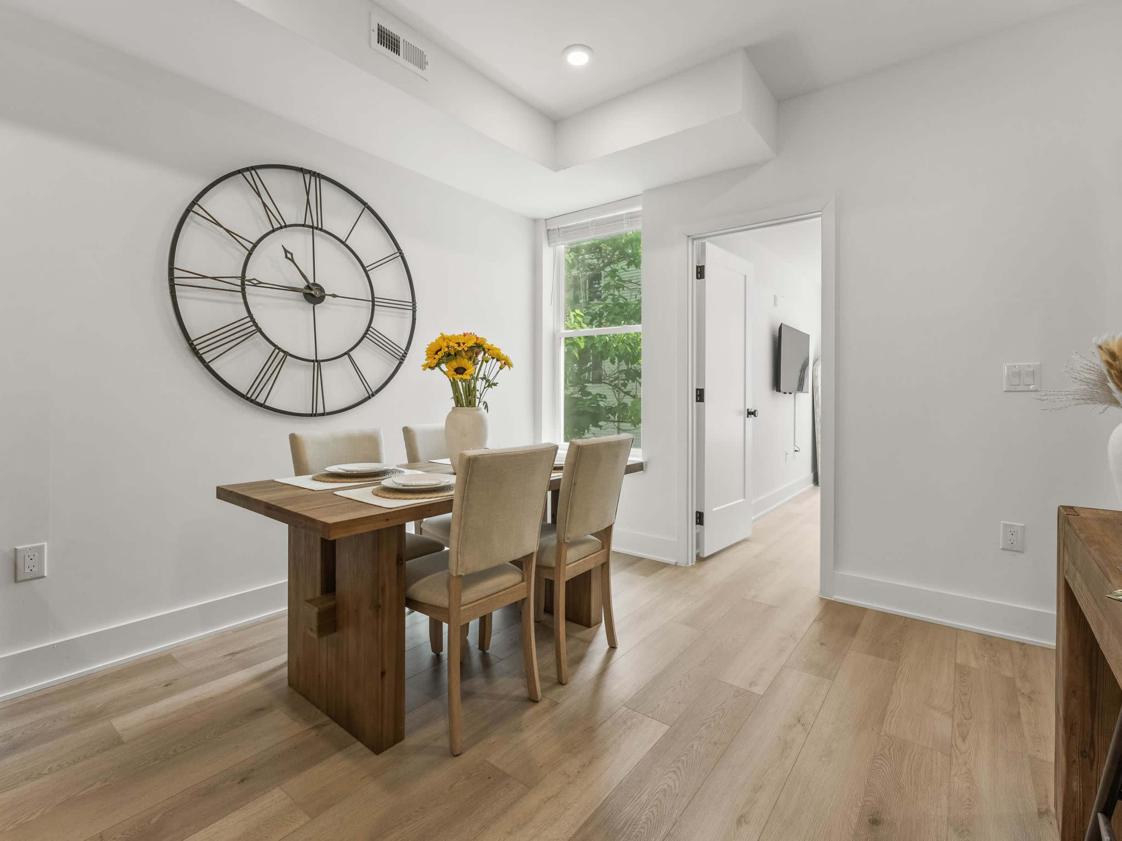 A dining area features a wooden table with four chairs, a large wall clock, and a vase of sunflowers on the table.