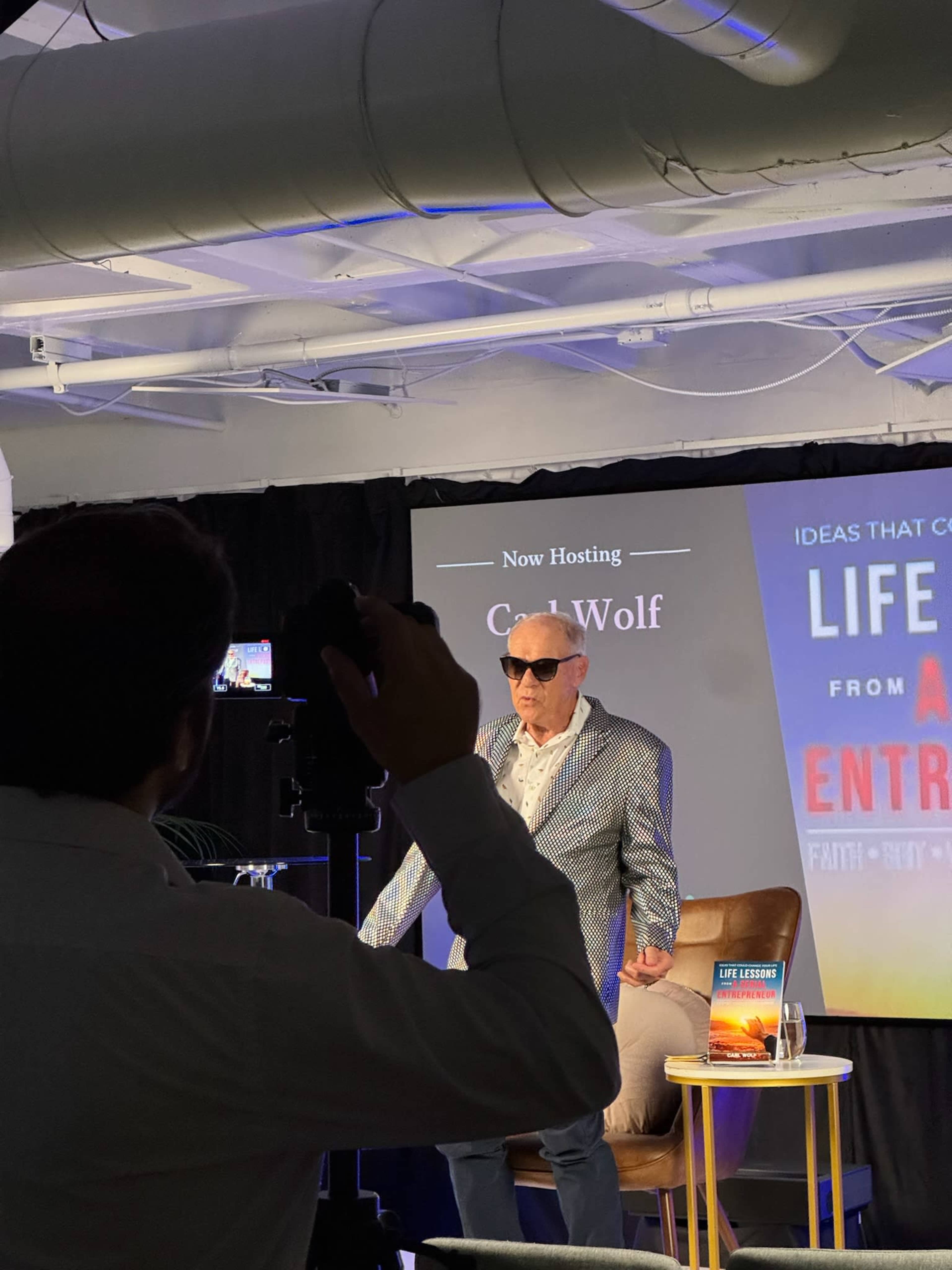 A man wearing sunglasses and a patterned jacket stands on stage in front of a banner that reads "Now Hosting Carl Wolf."
