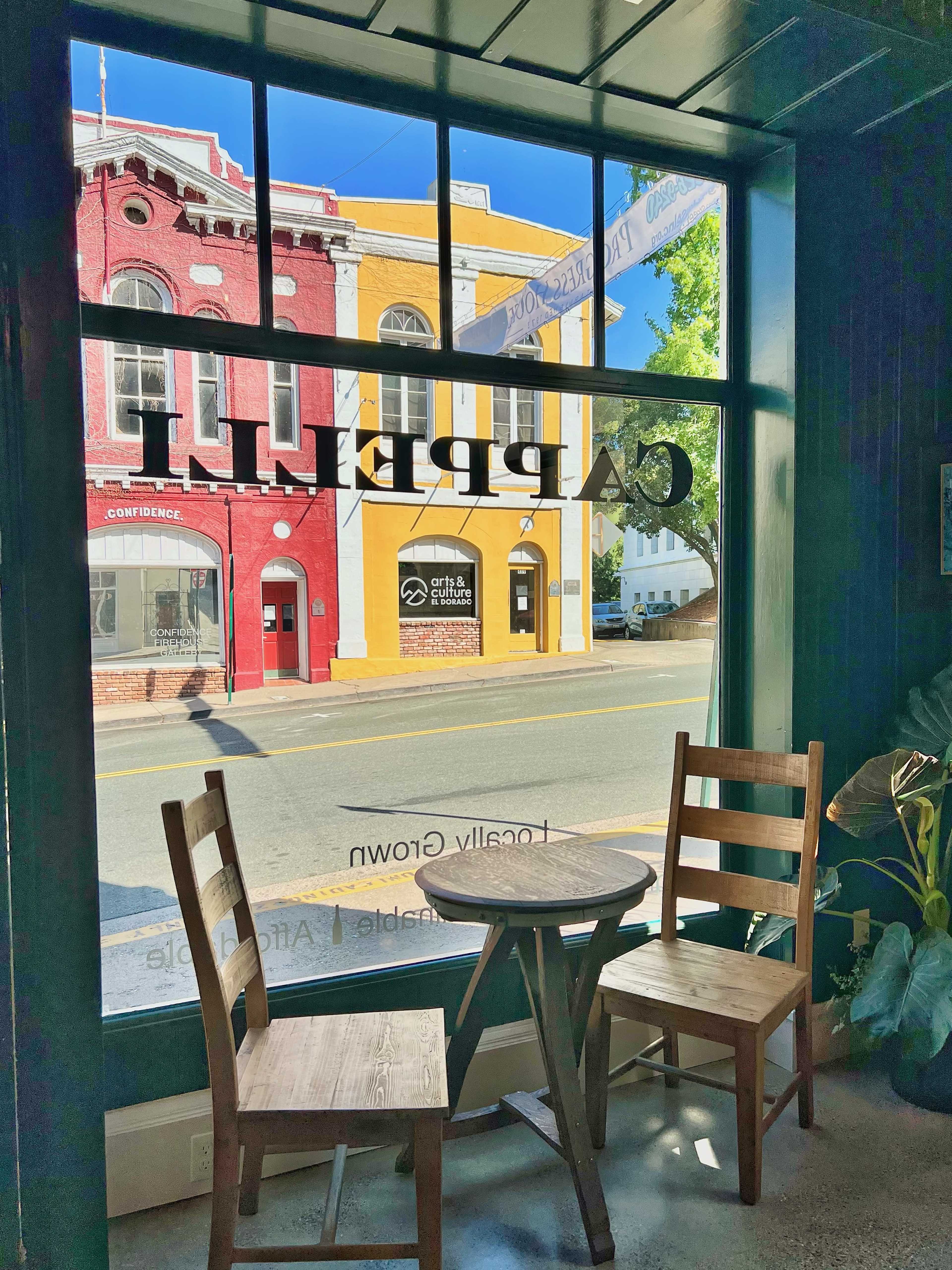 The image shows a view through a green-tinted window, featuring two wooden chairs and a small table, with colorful buildings visible across the street.