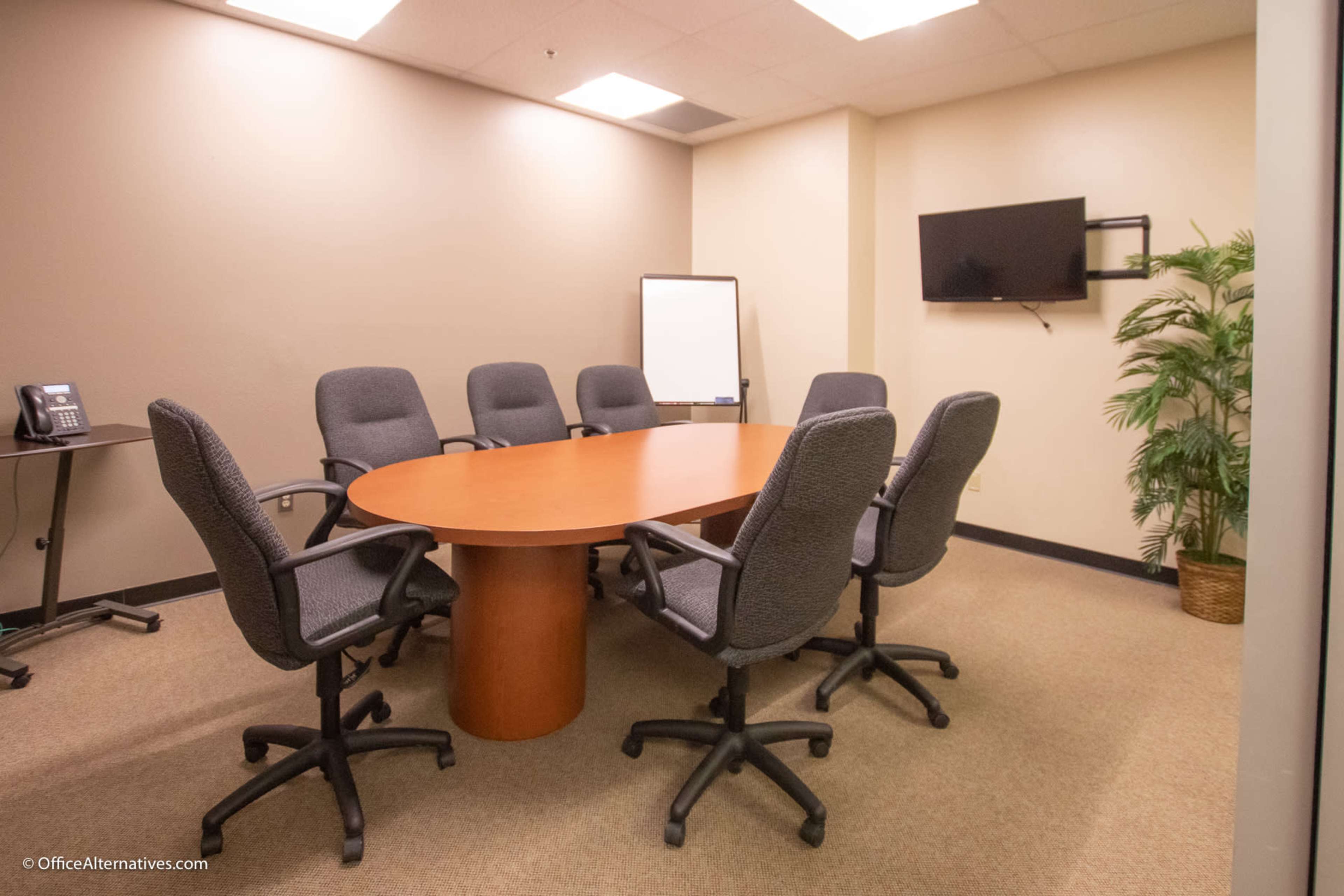 A conference room with a large oval table surrounded by eight office chairs, equipped with a whiteboard and a television.