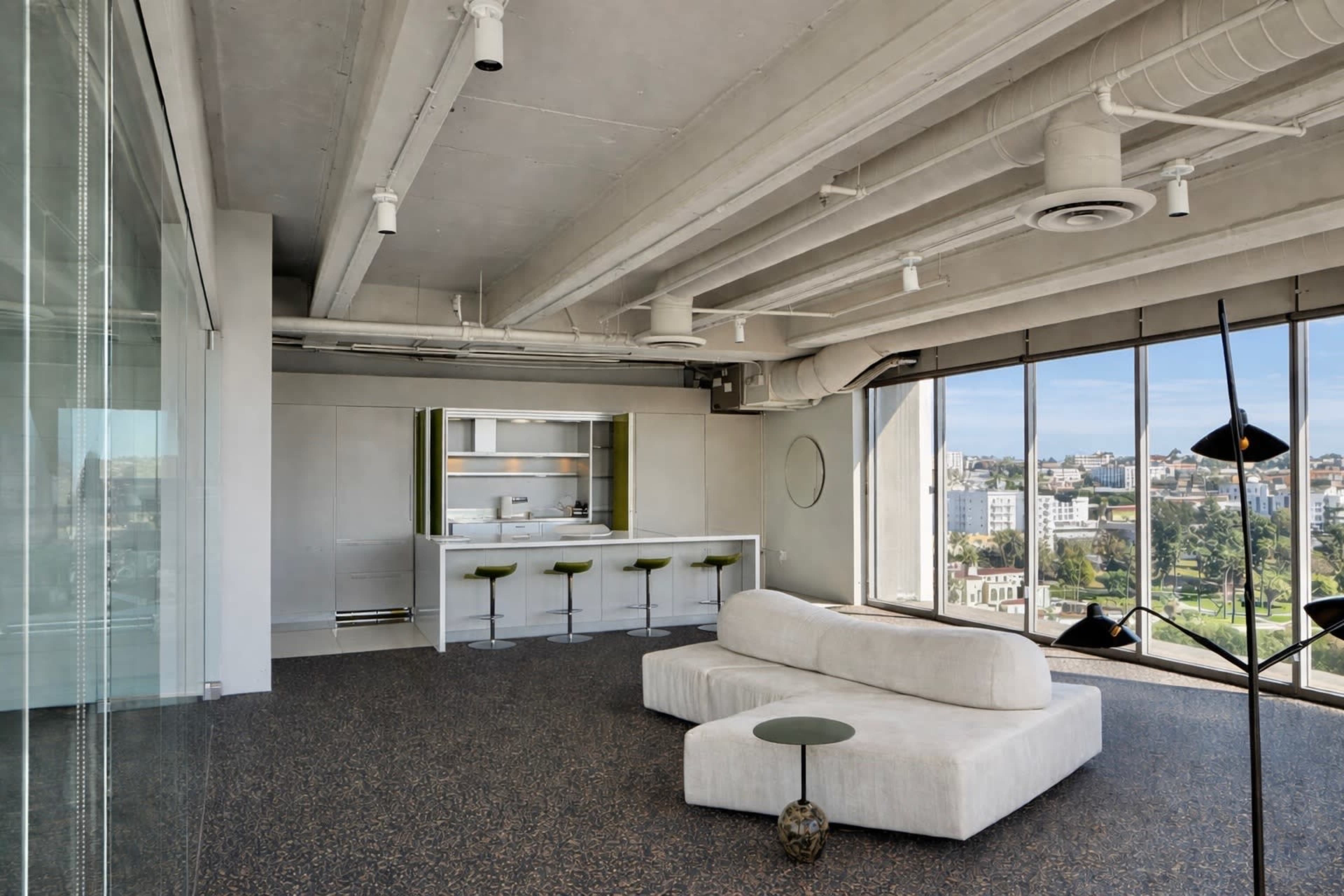 A modern living space features a minimalist design with a white sofa, a small green side table, and a kitchen area visible in the background, surrounded by large windows that offer a view of the outside.