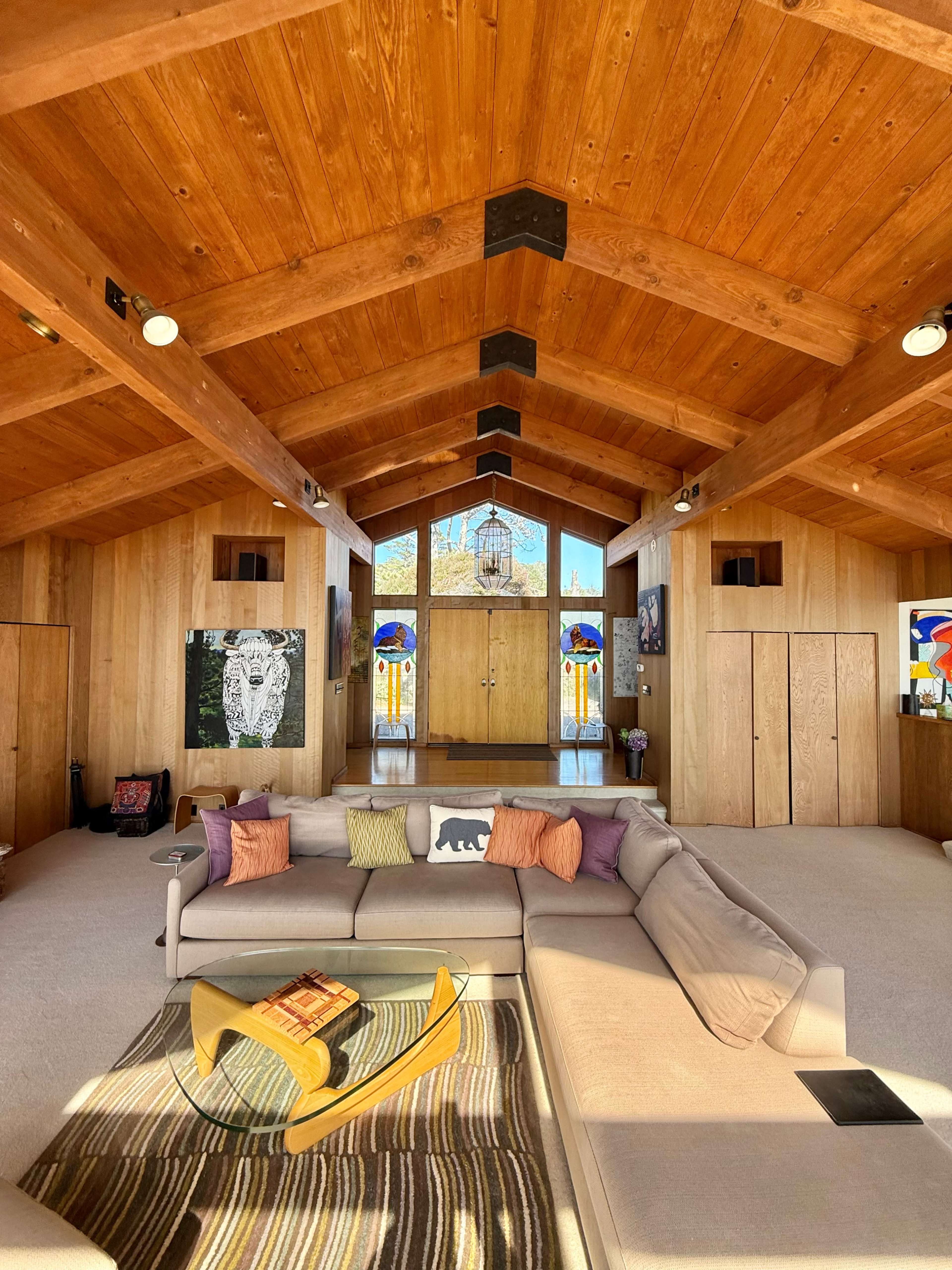 The interior of a wooden-beamed house features a spacious living area with a light-colored sofa, colorful throw pillows, and artwork on the walls, leading to large windows that showcase an outdoor view.