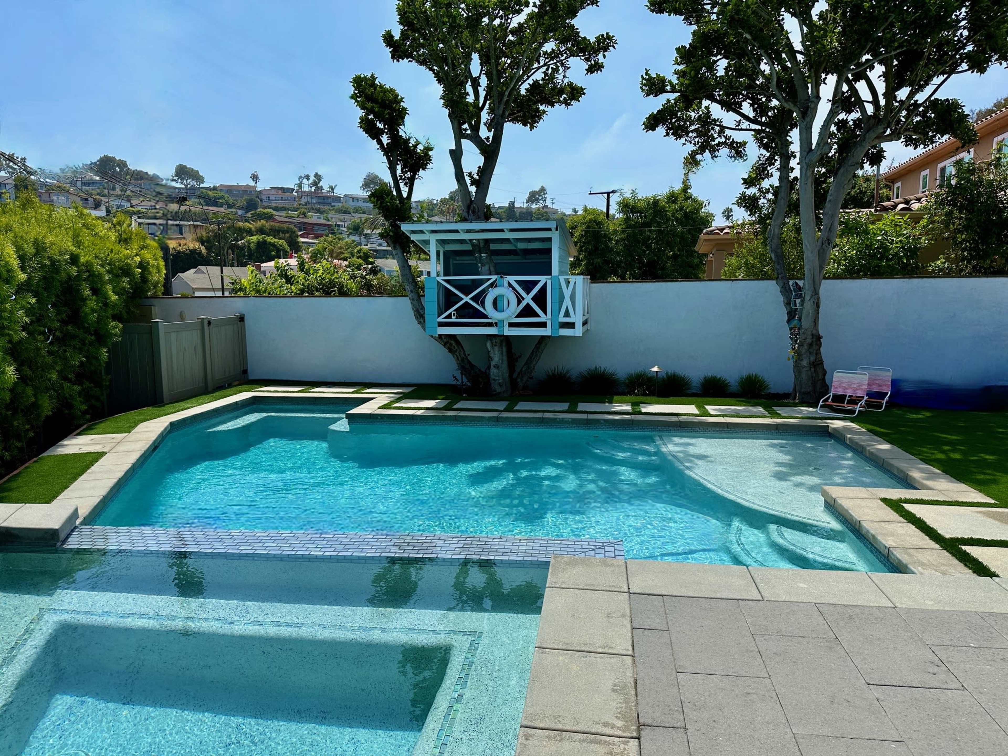 The image features a clear swimming pool surrounded by a well-maintained yard and a small lookout structure against a backdrop of hillside homes.