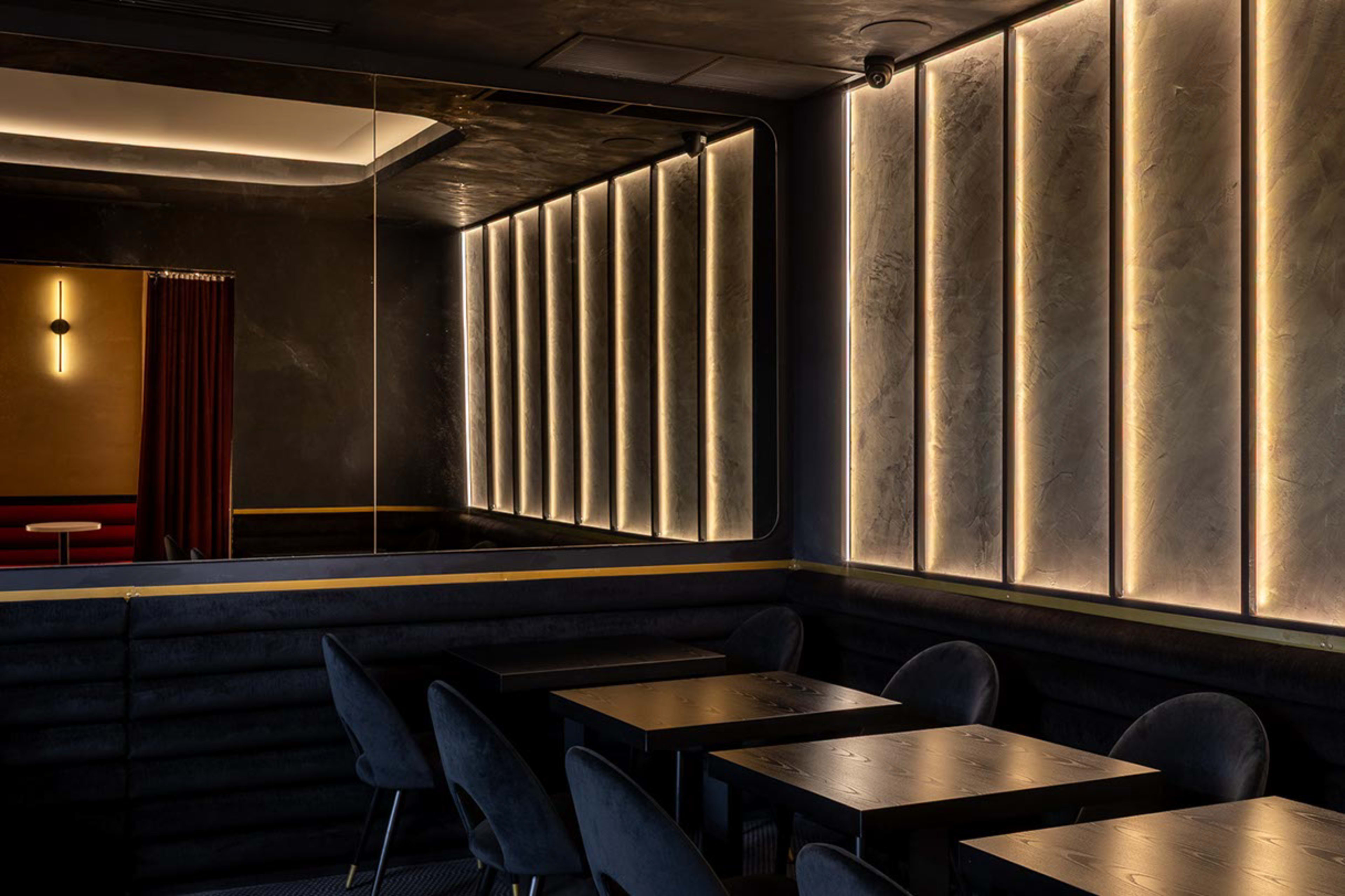 The image shows a dimly lit interior of a lounge featuring dark walls, illuminated vertical panels, and several tables with upholstered chairs.