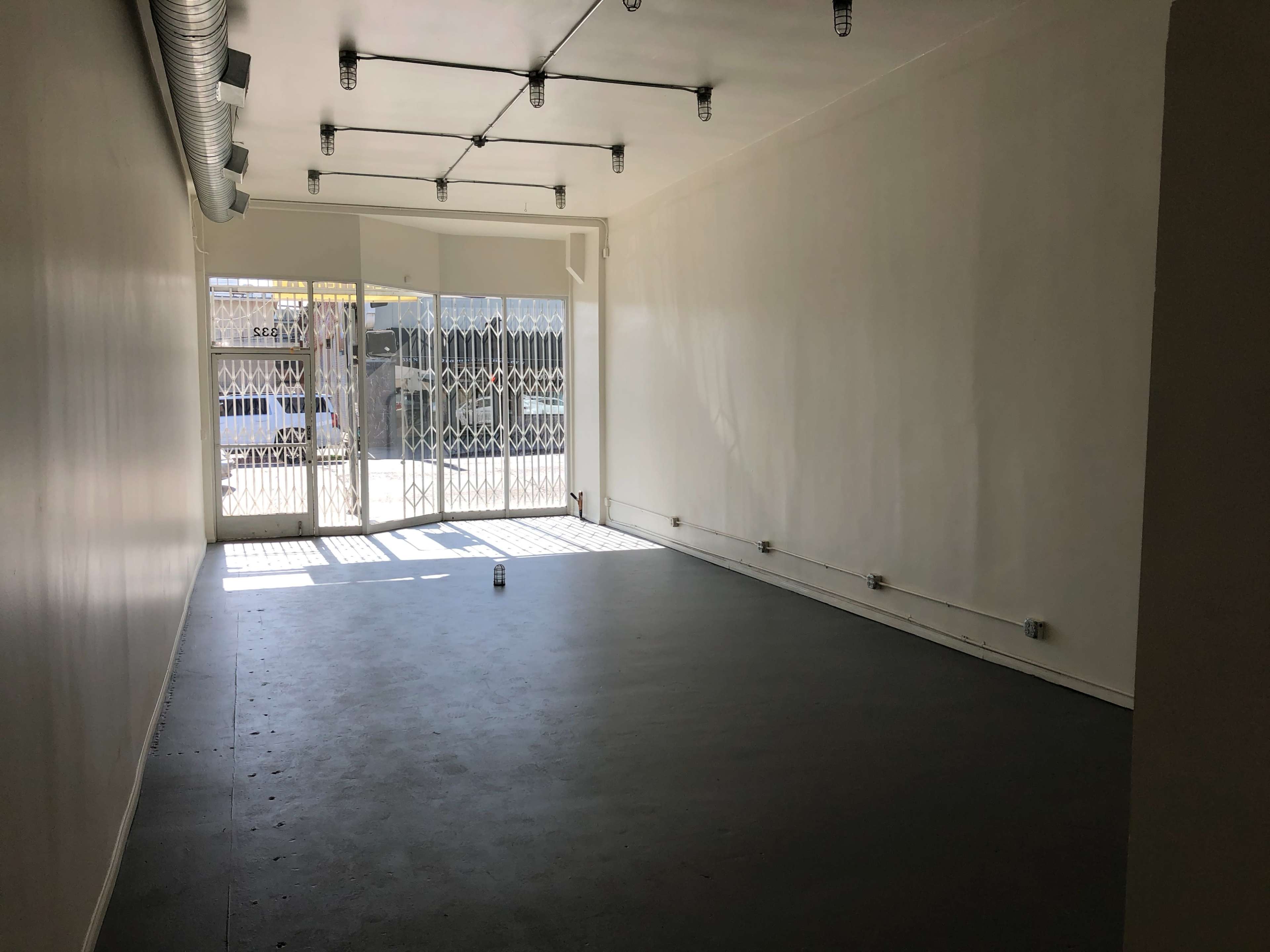 The image shows an empty room with a concrete floor and large windows covered by a security grille, allowing light to enter.