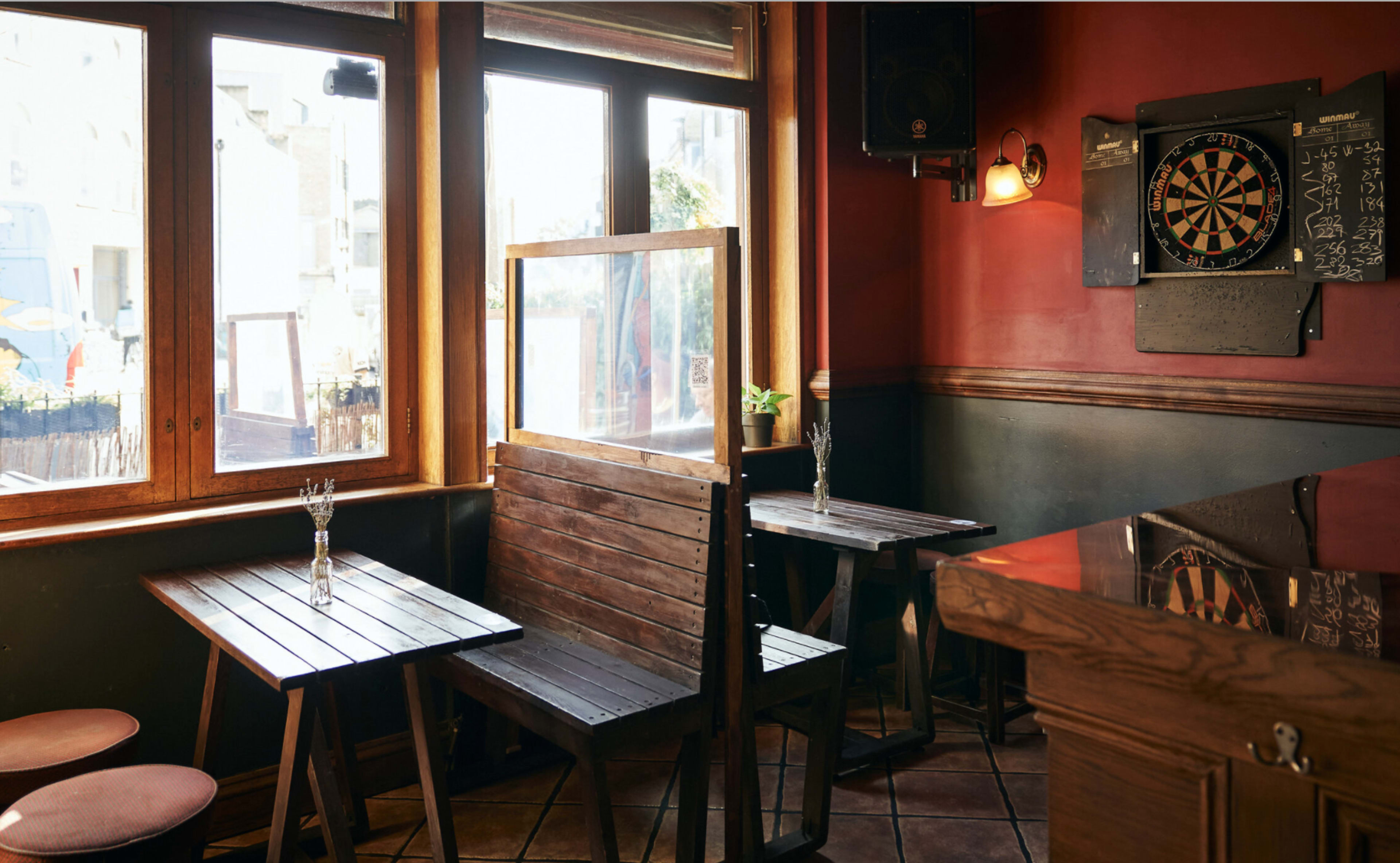 A cozy pub interior with wooden tables, a dartboard on the wall, and large windows allowing natural light.