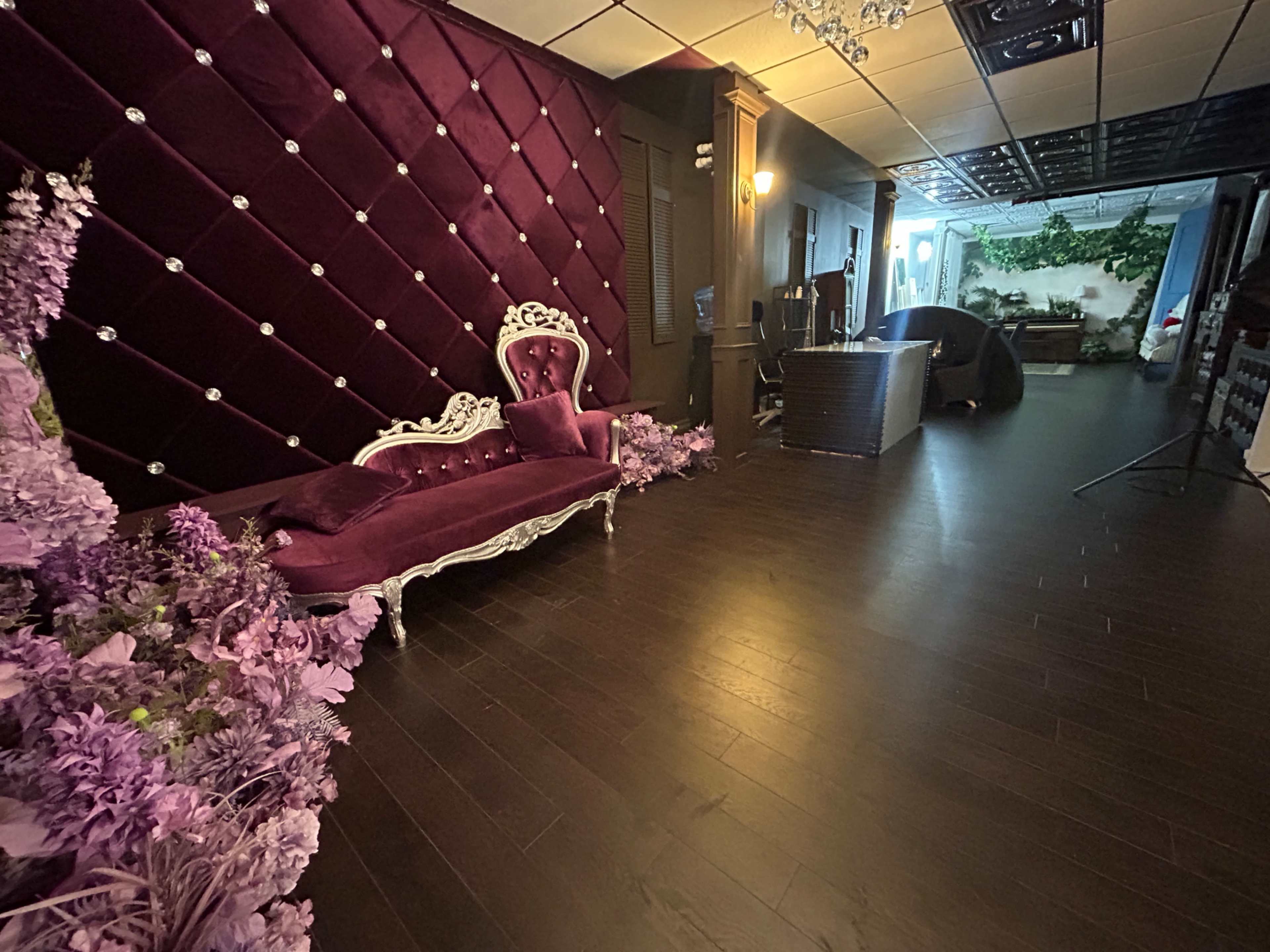 The image depicts an interior space featuring a purple velvet tufted wall, an ornate white sofa, and floral decorations lining the floor.