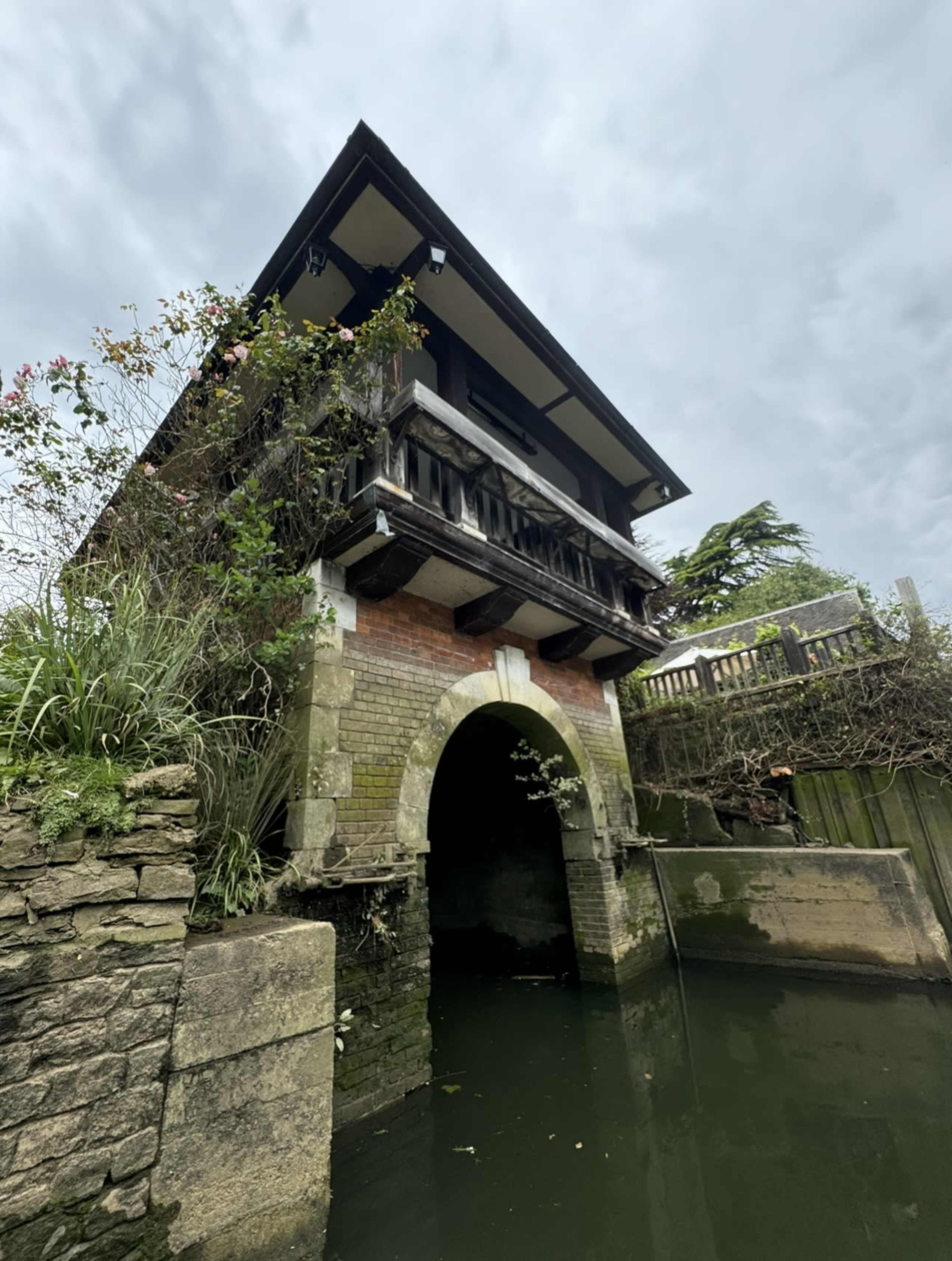 A structure with a wooden balcony and brick facade sits atop an archway over water, surrounded by greenery and cloudy skies.