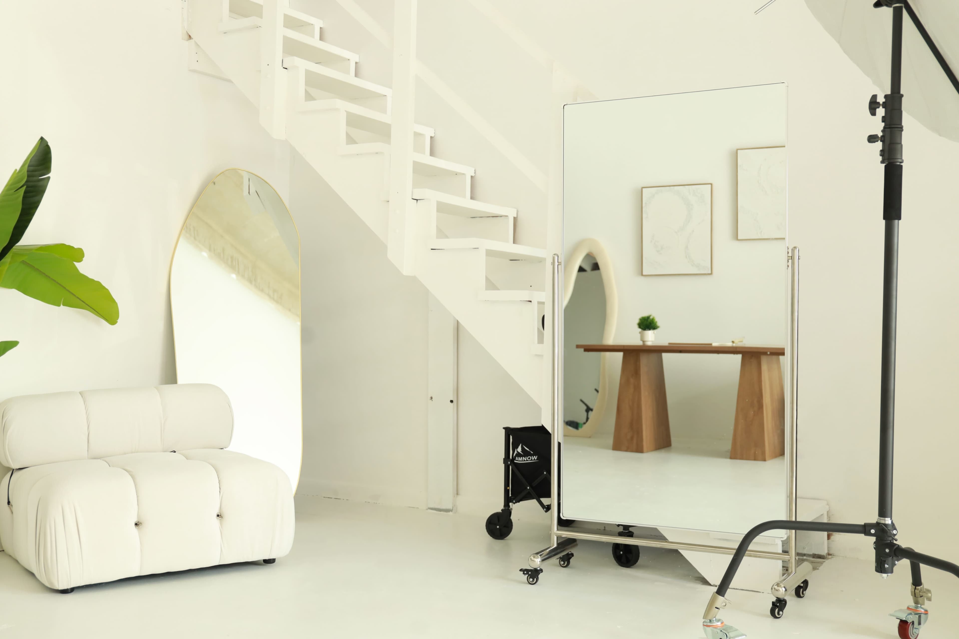 The image shows a bright, minimalistic interior featuring a staircase, a large mirror, a light-colored sofa, and a wooden table with a small decorative plant.