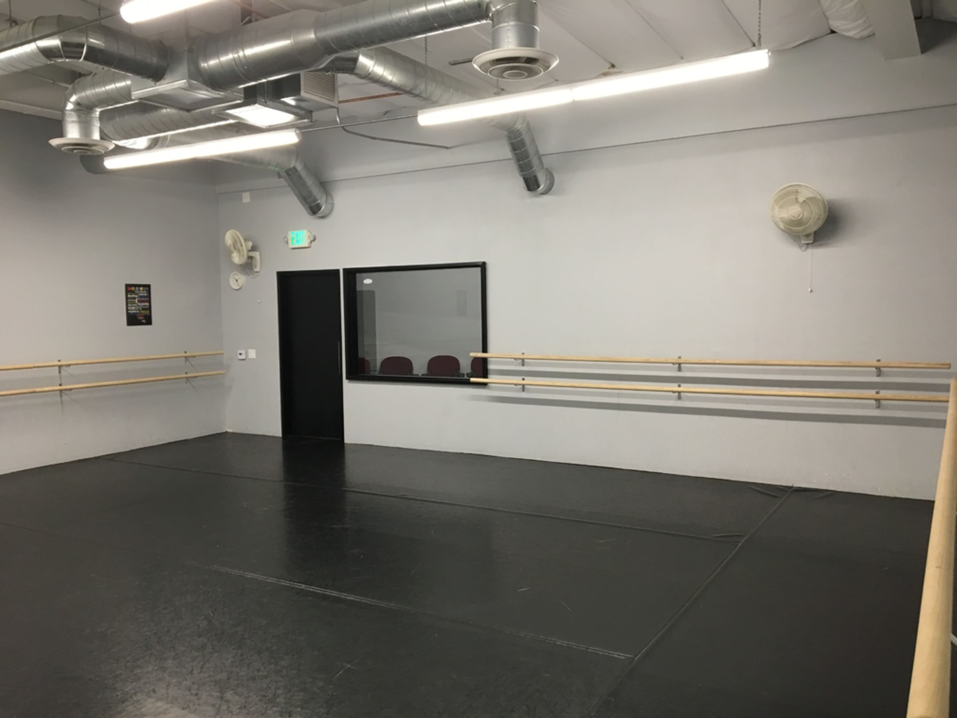 An empty dance studio with a black floor, mirrored wall, and ballet barres along two sides.