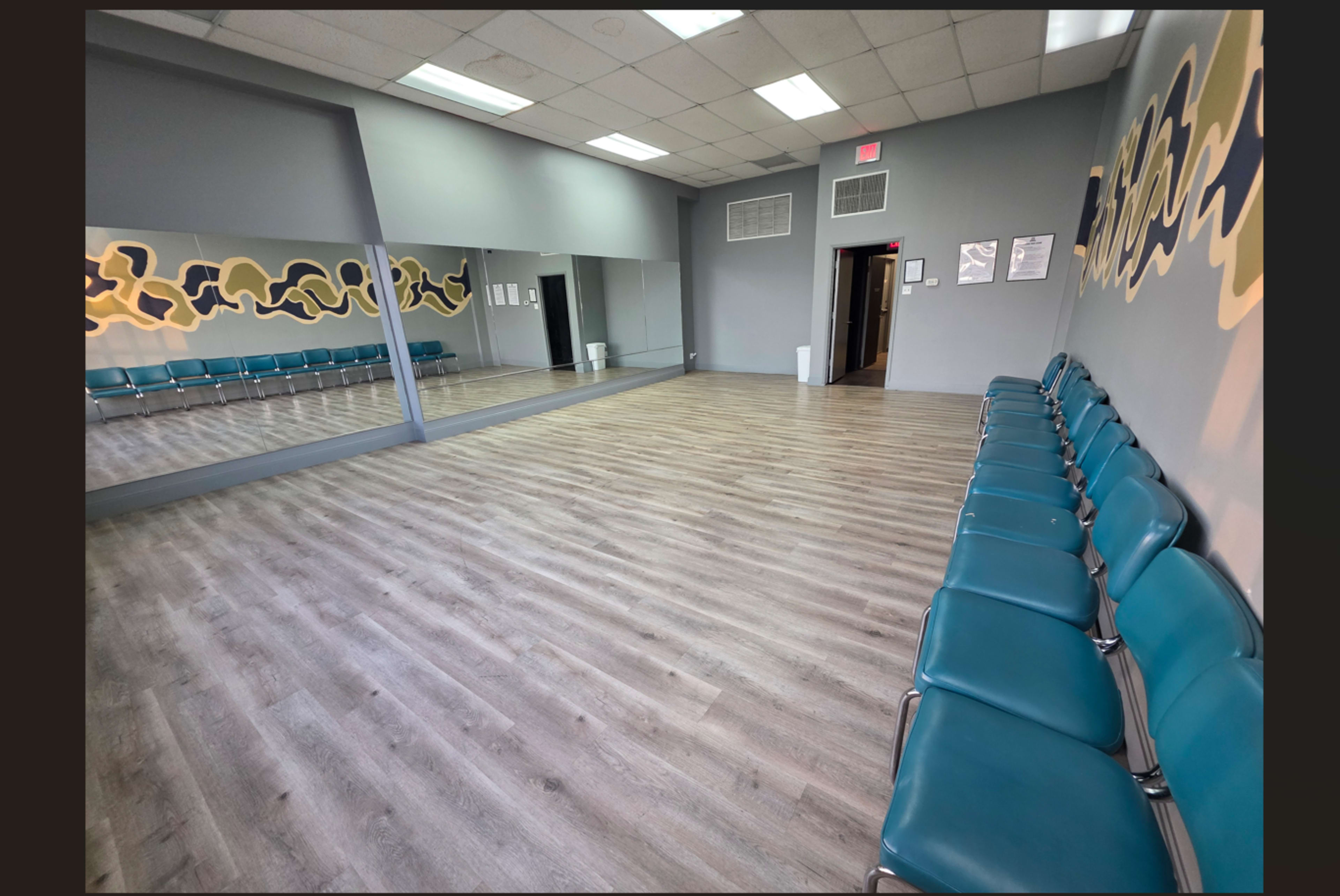 The image shows a spacious waiting room with teal chairs arranged along one wall and a large mirror on the opposite wall.