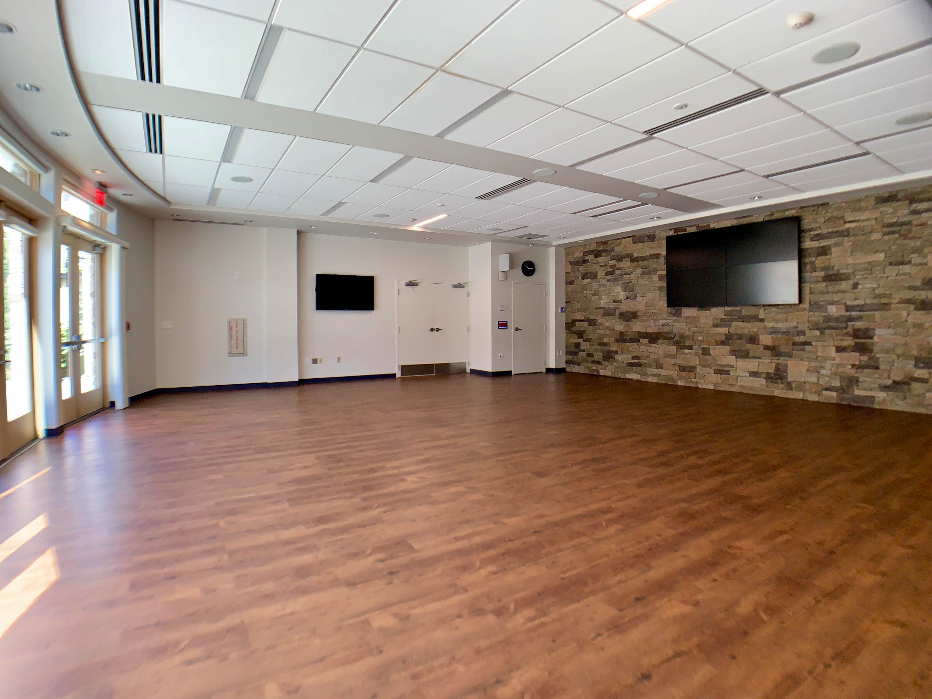 A spacious, empty room with wooden flooring, large windows, and a stone wall featuring a mounted television.