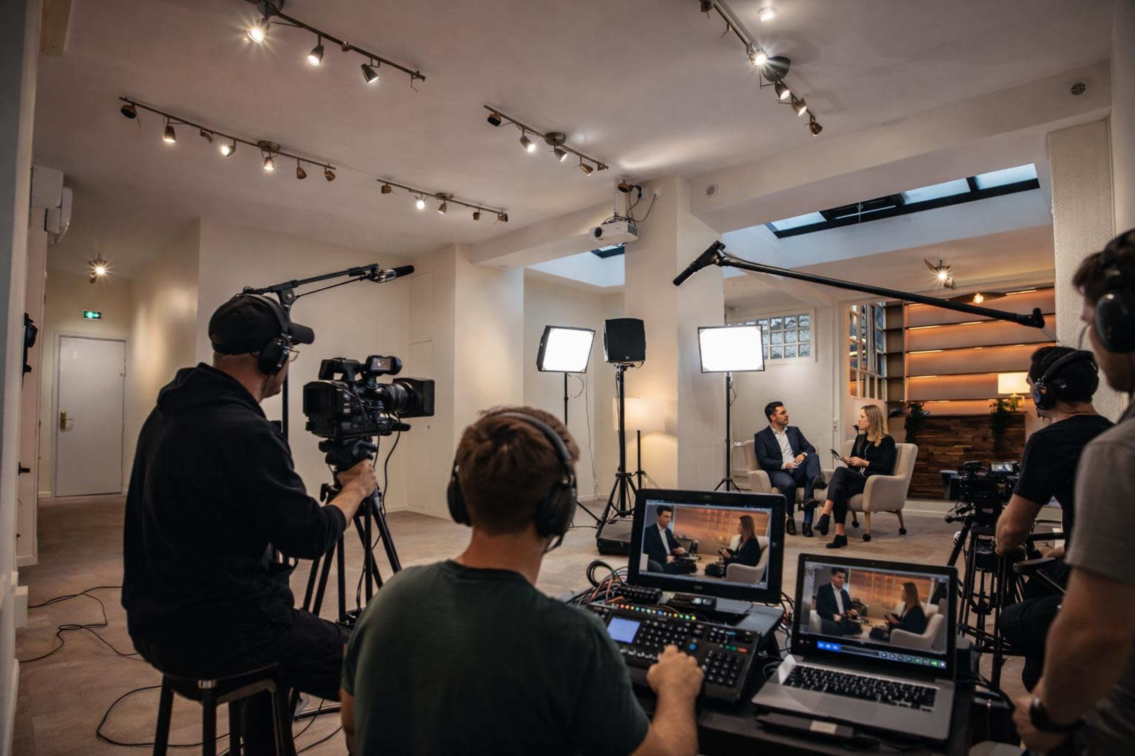 A television interview is being filmed in a well-lit studio with multiple cameras and crew members.