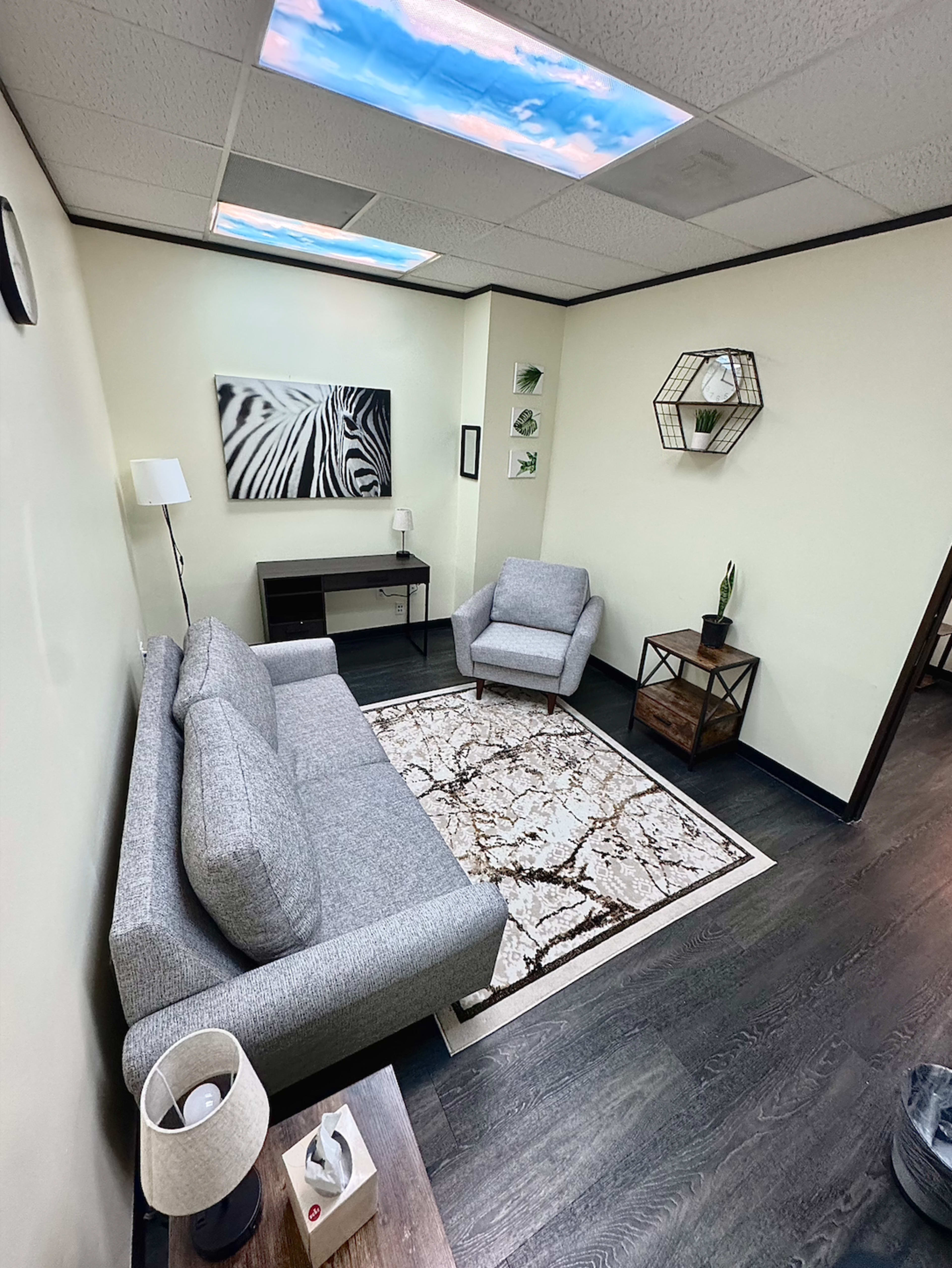 The image shows a small, modern waiting room with a gray sofa, an armchair, a coffee table, and decor on the walls.