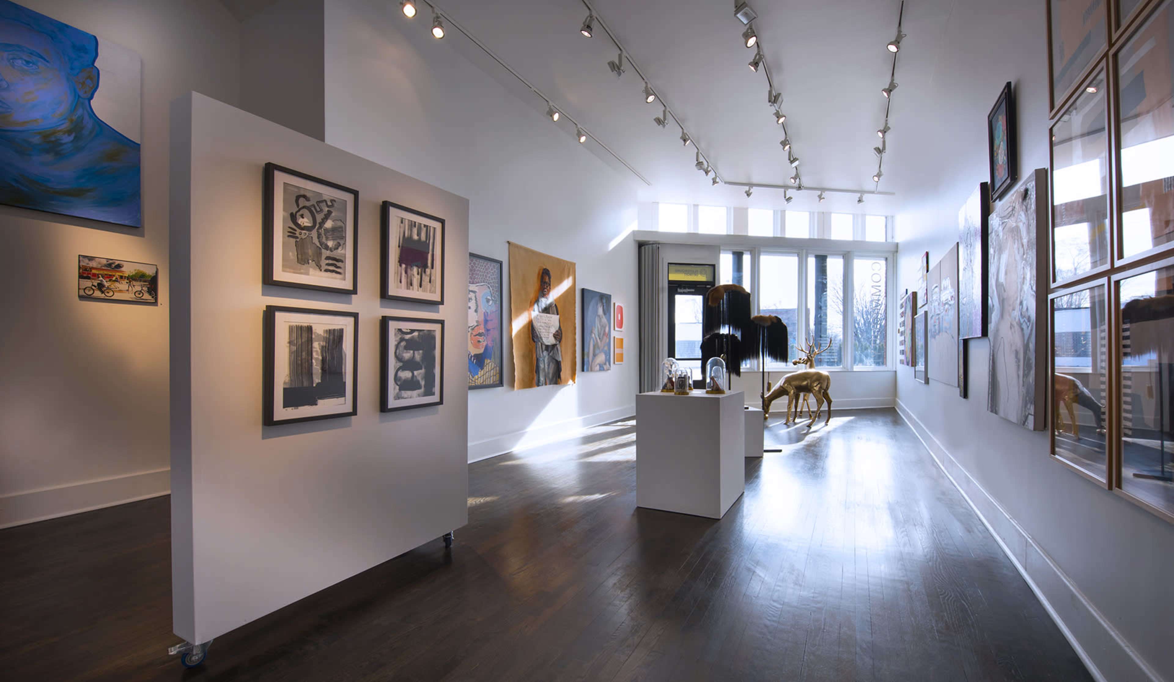 The image shows an art gallery with white walls, wooden flooring, and various artworks displayed in frames and on pedestals.