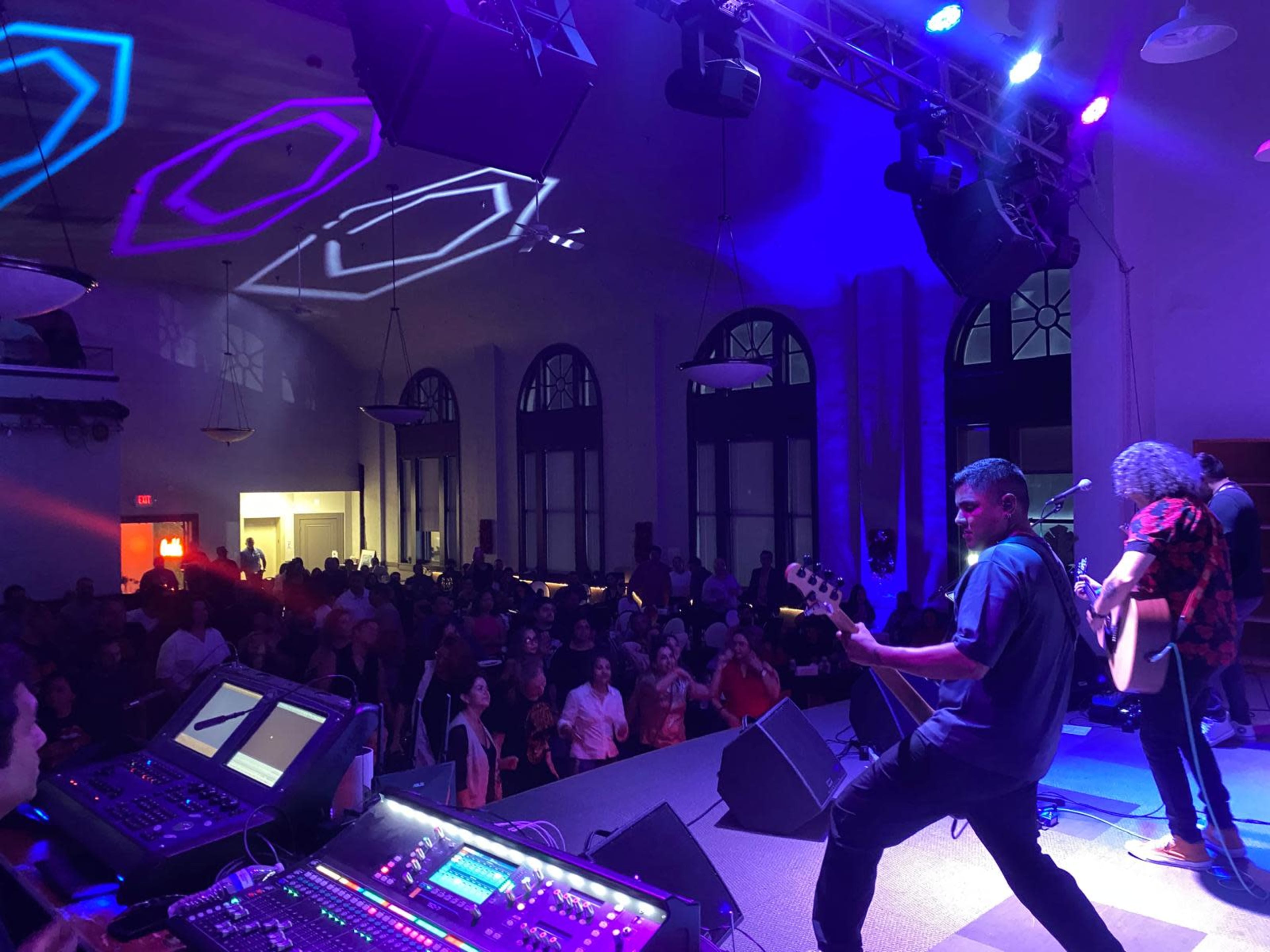 A band performs on stage in front of a lively audience in a dimly lit venue with colorful lights and sound equipment visible in the foreground.